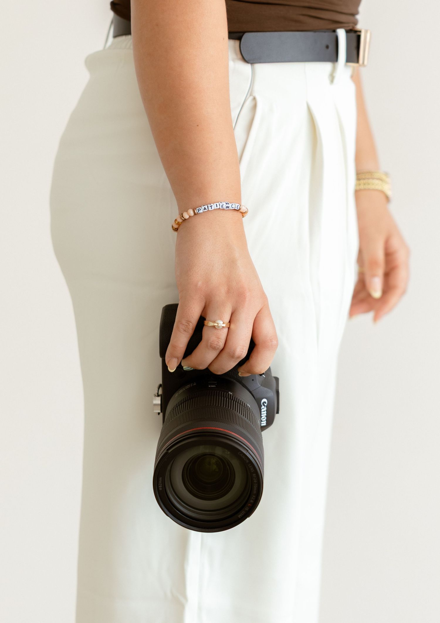 Woman in white trousers holding a Canon DSLR camera with a telephoto lens, wearing gold bracelets and a ring.