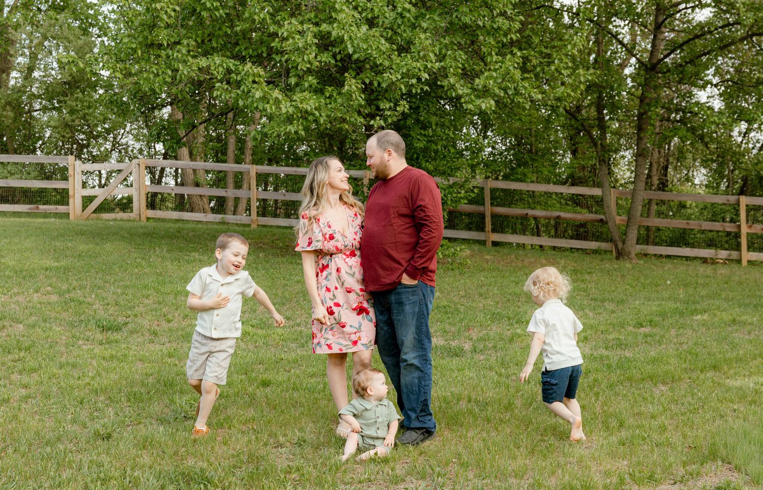 Family of five posing outdoors on green grass with wooden fence, parents smiling while three young children play around them.
