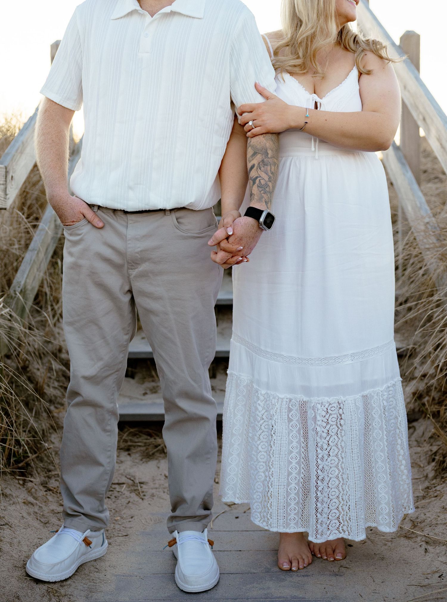 Couple holding hands on a beach boardwalk, man in khaki pants and white shirt, woman in white lace maxi dress.