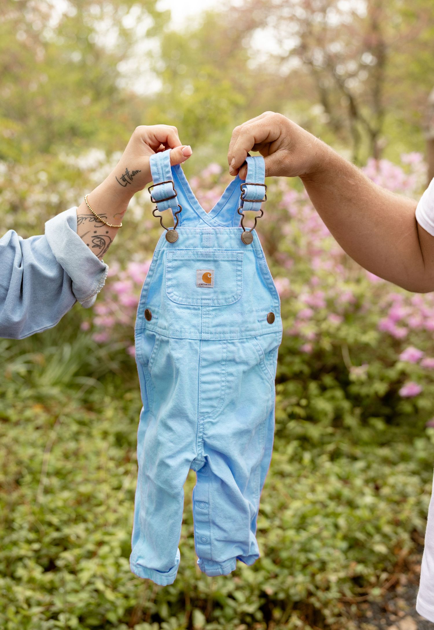 Two hands holding tiny light blue Carhartt baby overalls outdoors with pink flowering bushes in background.