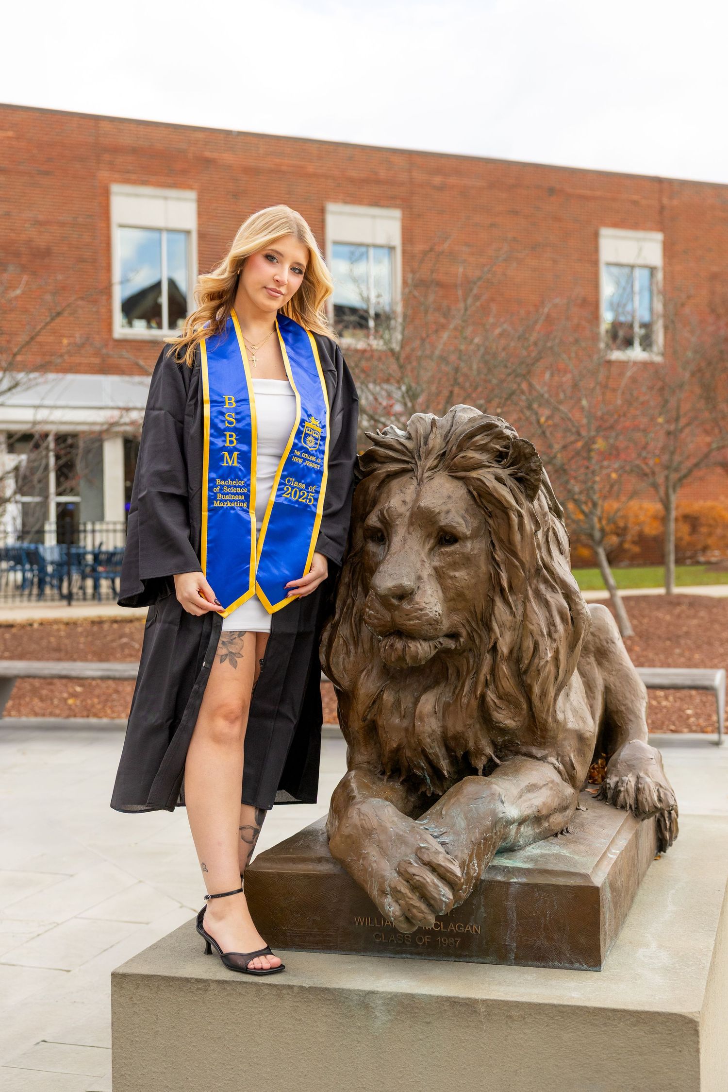 Graduate in academic regalia poses next to large bronze lion statue outside brick campus building.