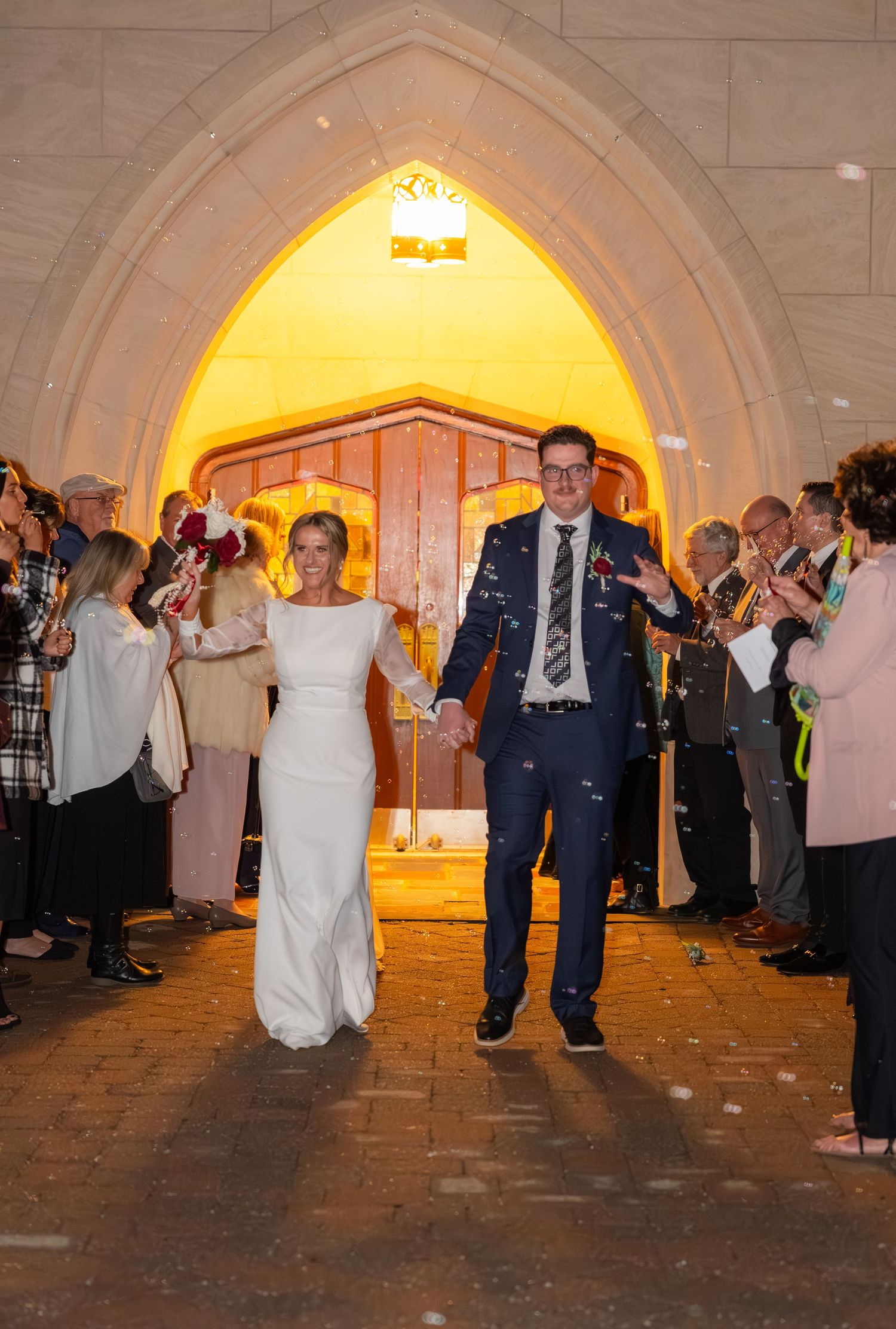 Bride and groom exit church through Gothic arch as guests toss confetti during evening wedding celebration.