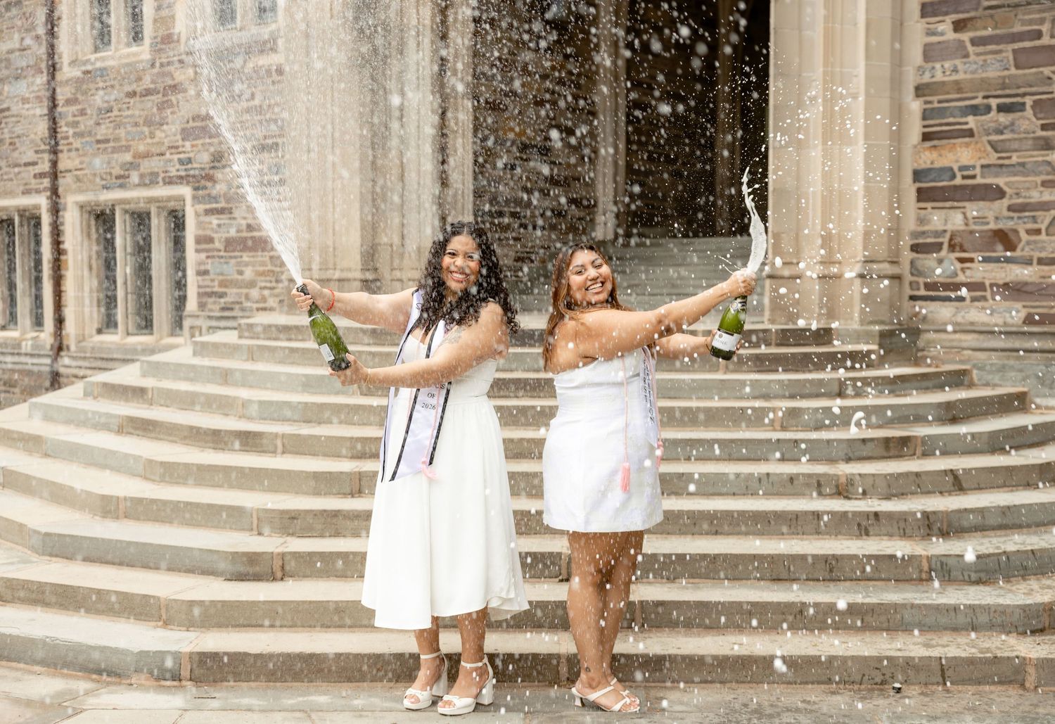 Two women in white dresses celebrate graduation by popping champagne bottles on stone steps.