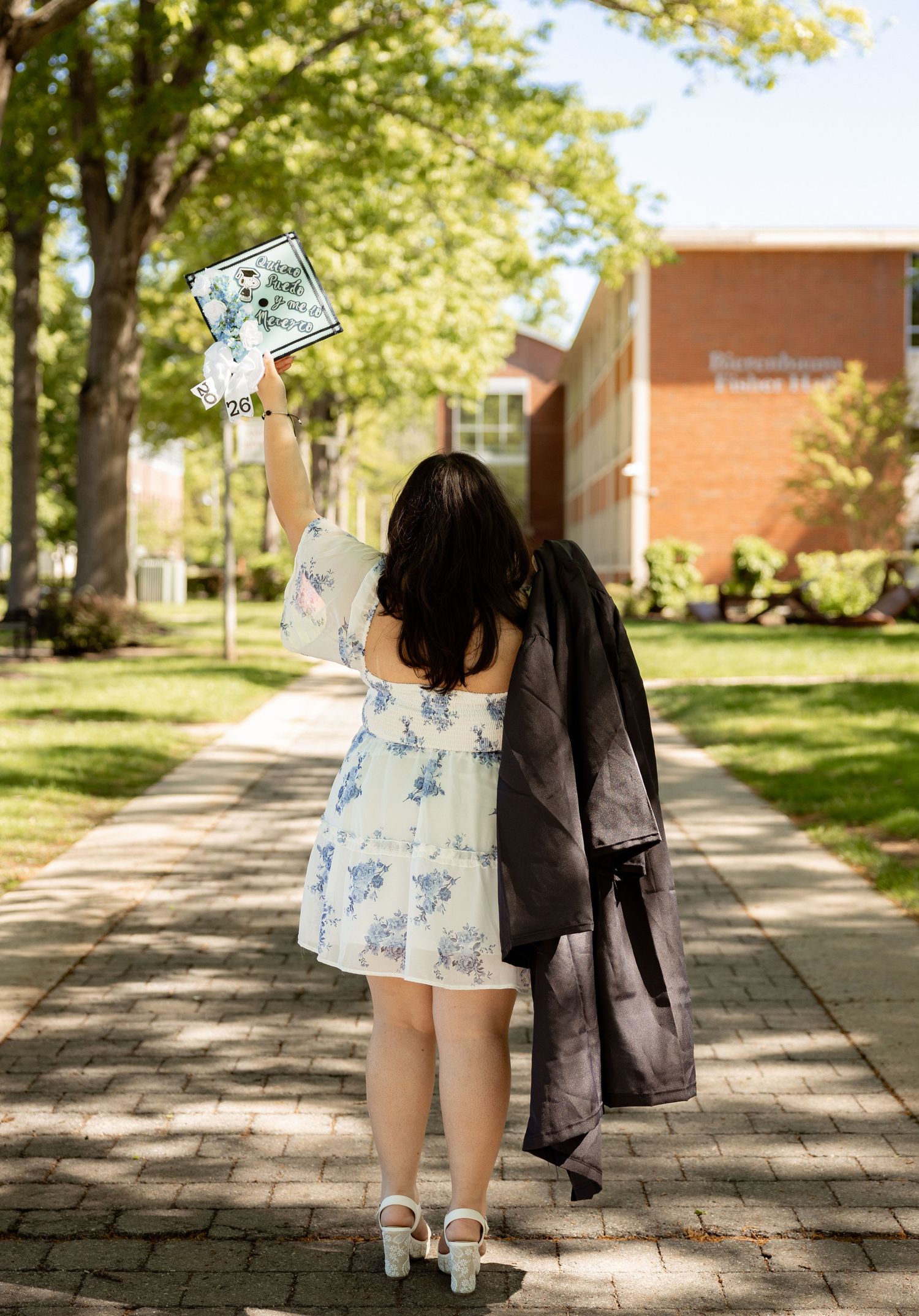 Graduate in floral dress raises decorated cap on brick campus path, holding gown, celebrating college graduation outdoors.