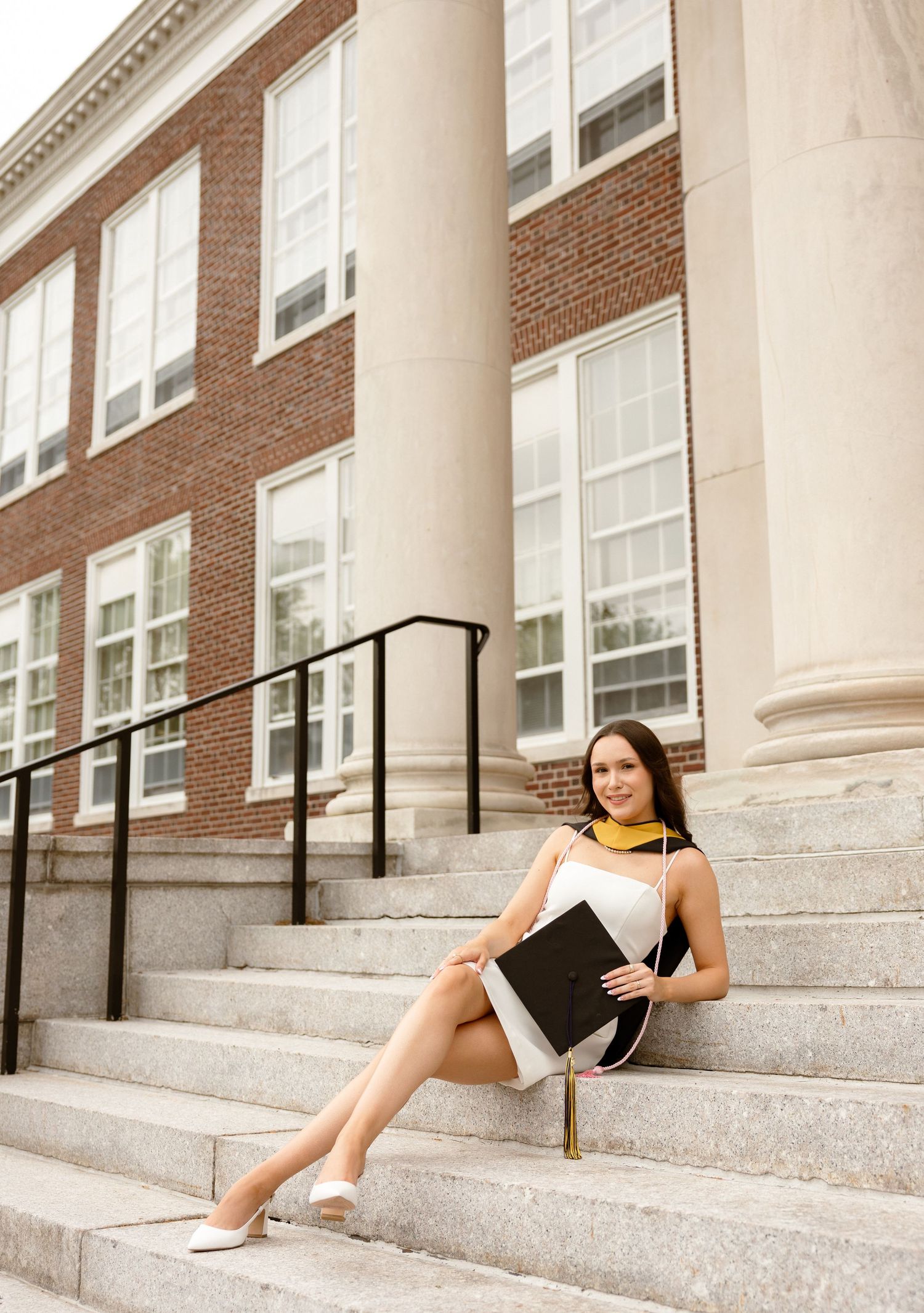 A young woman in white dress with graduation stole sits on university steps holding diploma cover.