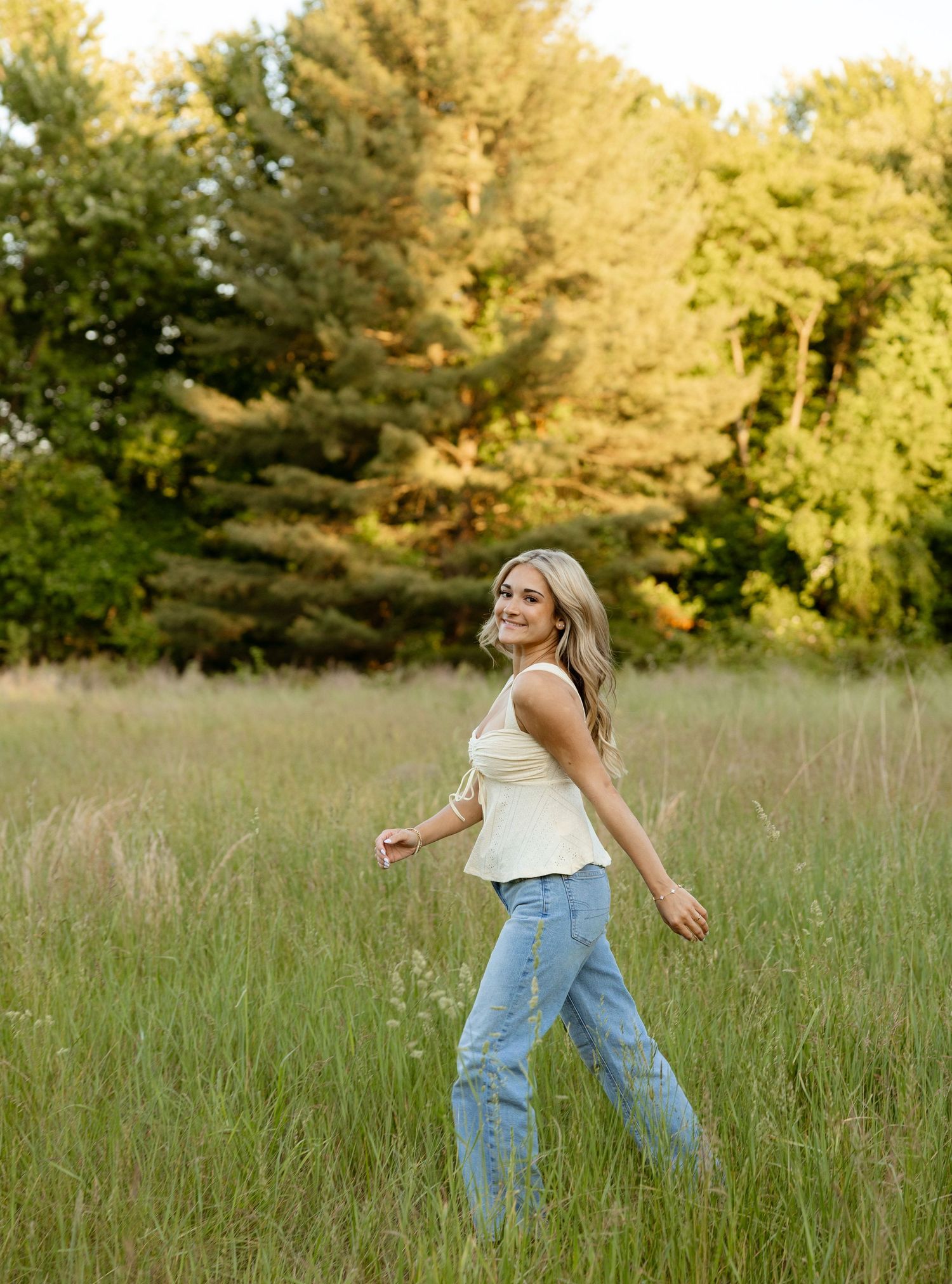 A smiling blonde woman walks through a lush green meadow wearing a white top and light blue jeans with trees behind her.