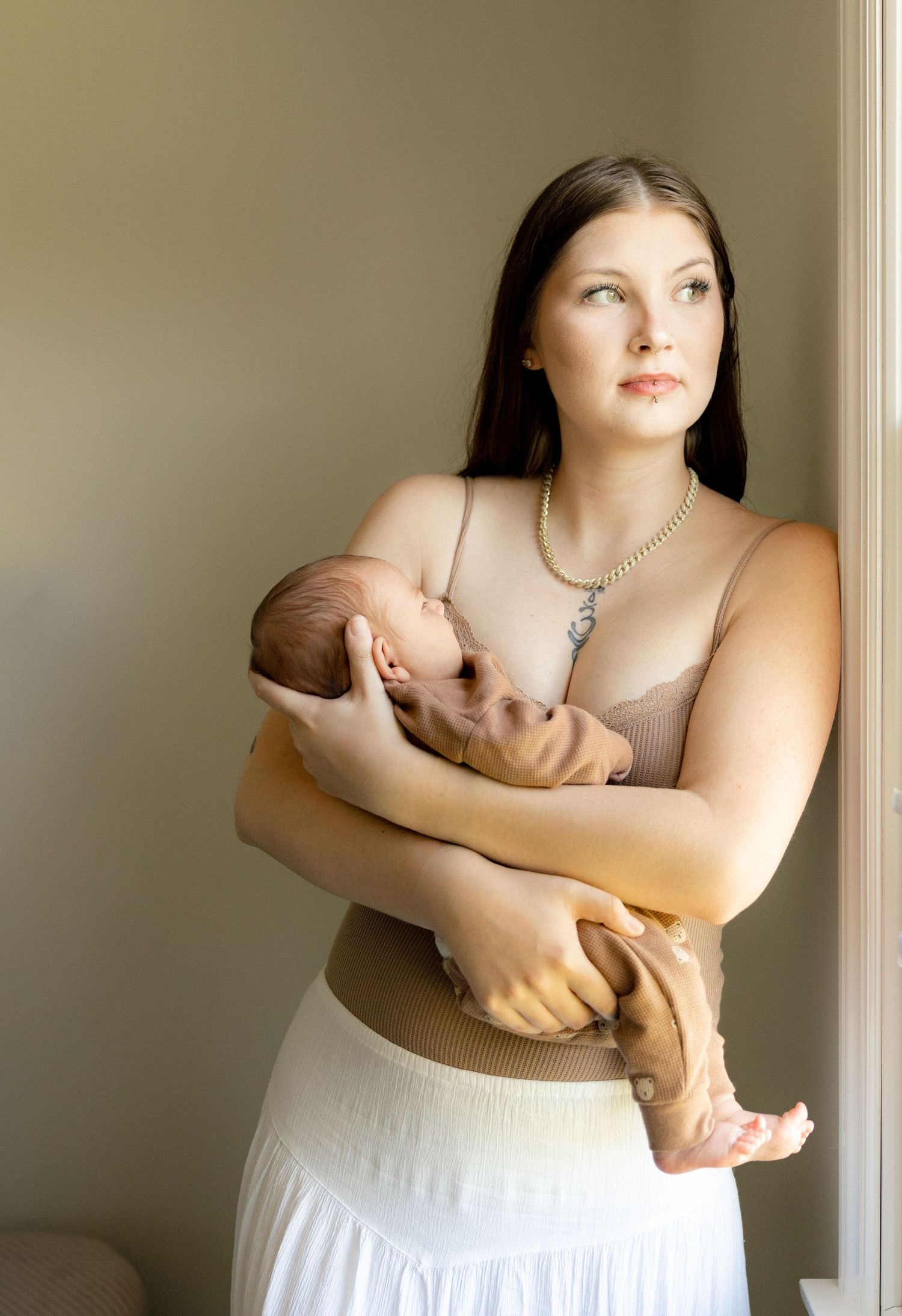 Young mother in beige top and white skirt tenderly holds sleeping newborn baby near a sunlit window.
