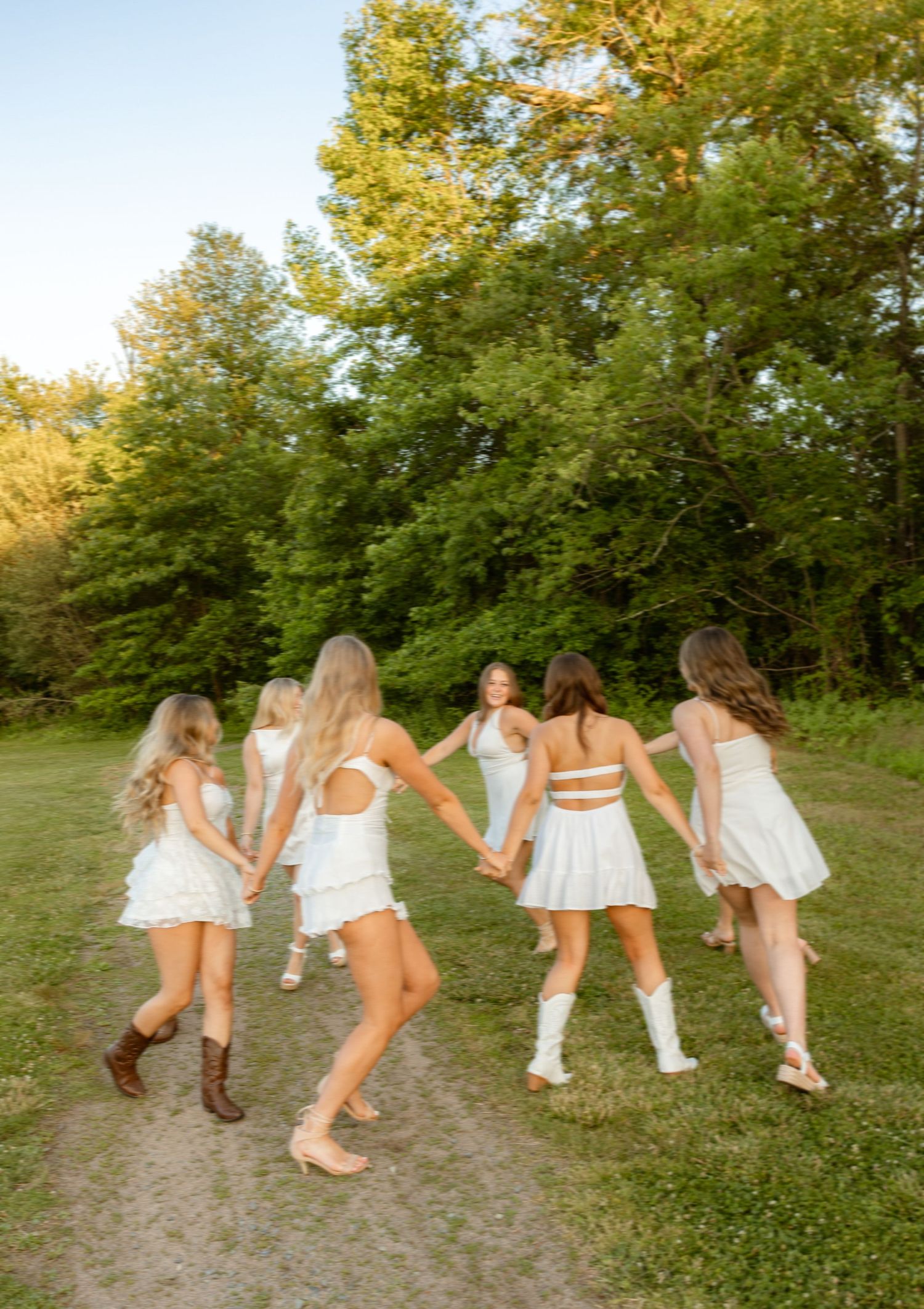Six young women in white dresses hold hands in a circle on a grassy path surrounded by lush green trees.
