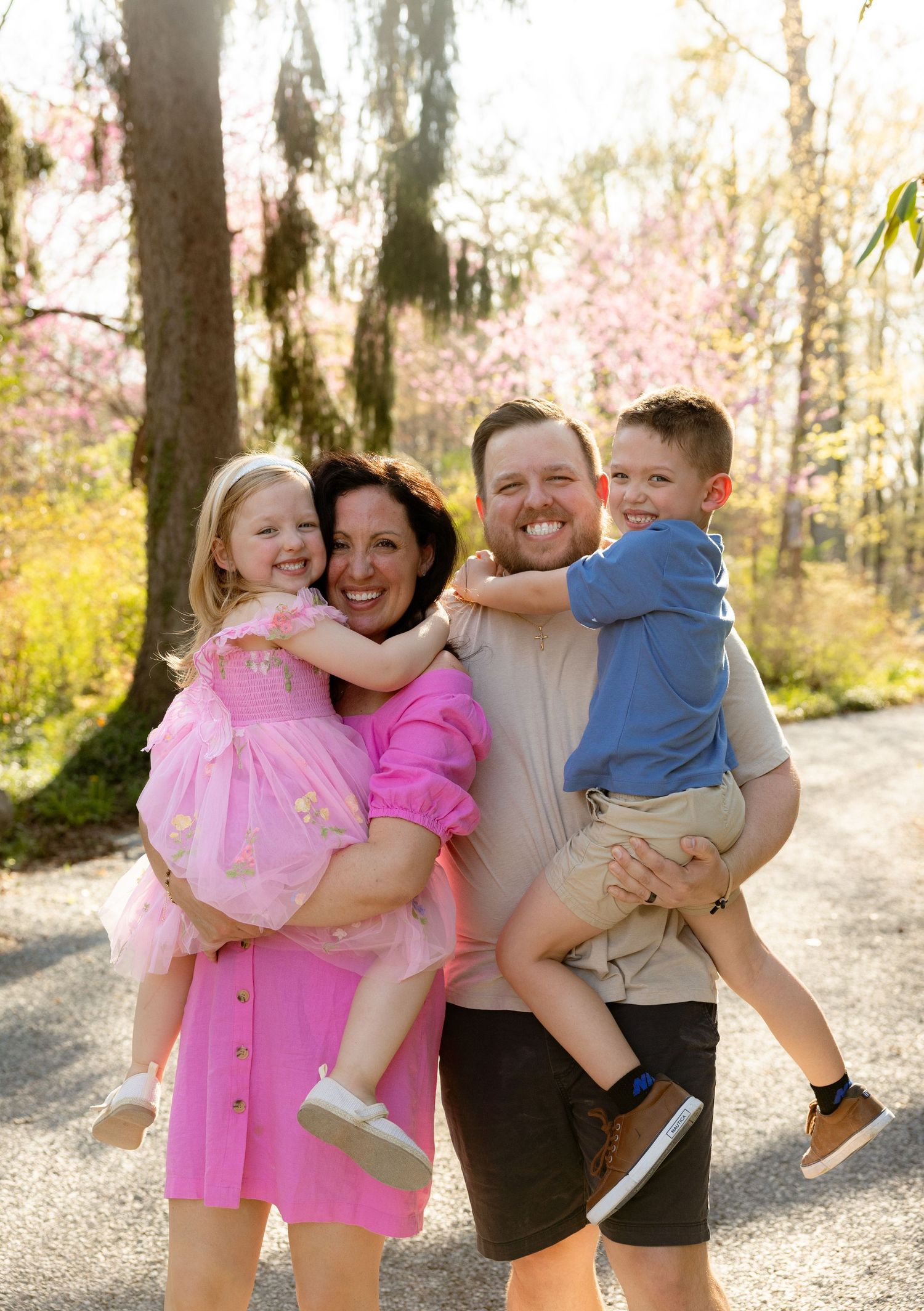 A happy family of four poses outdoors in spring, parents holding two young children among pink blooming trees.