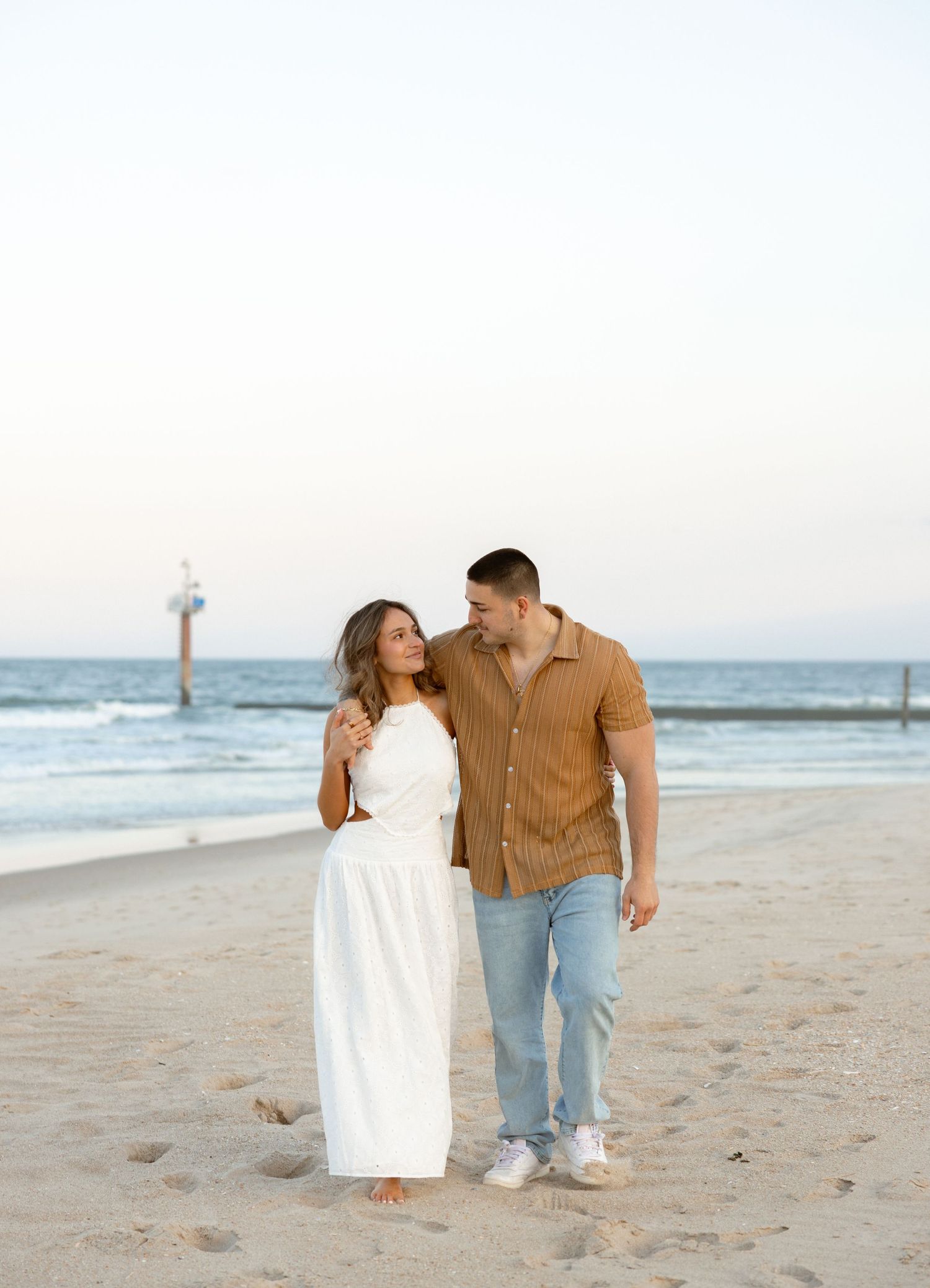Couple walking on beach, woman in white dress, man in brown shirt and jeans, ocean backdrop at dusk.