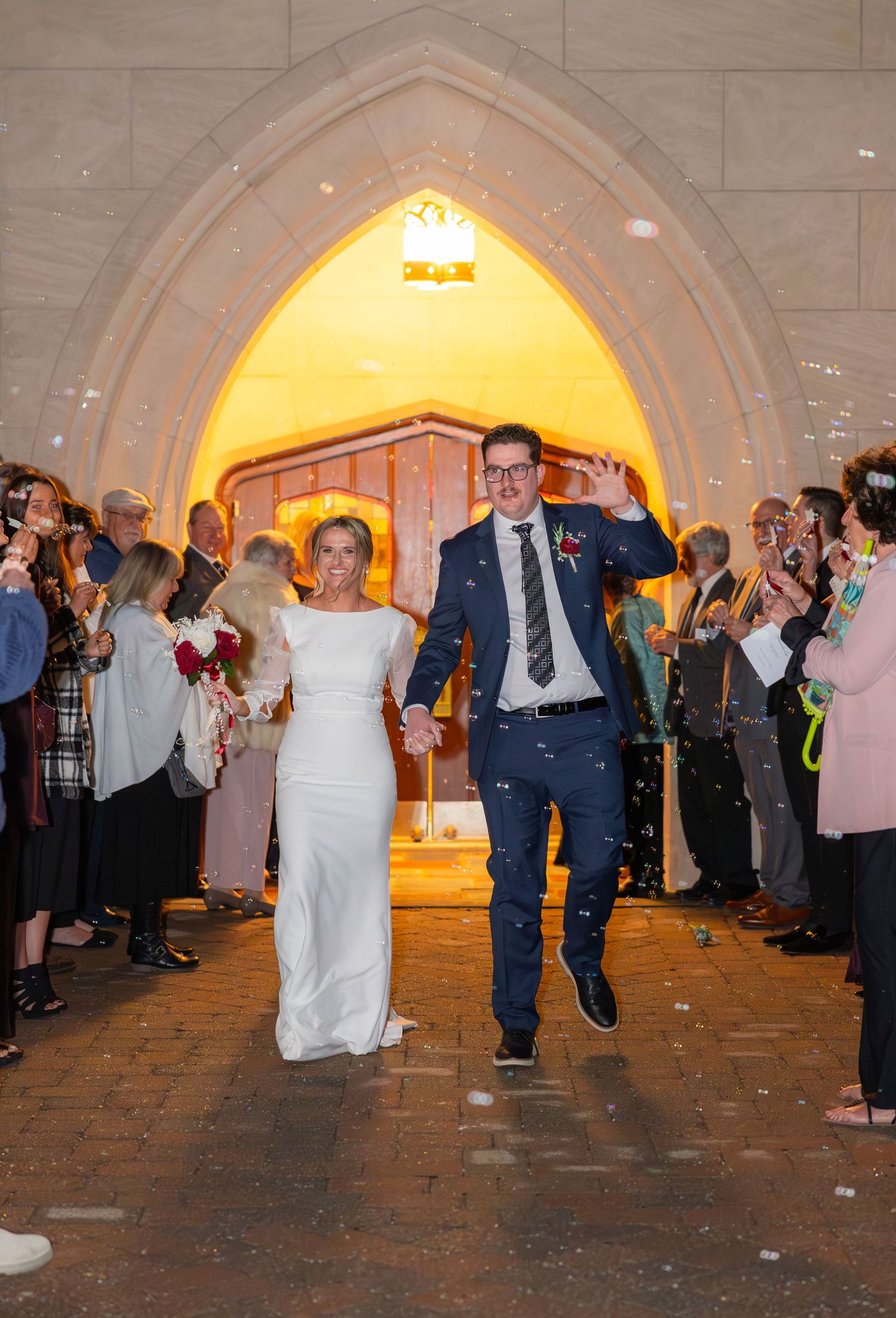 Newlywed couple makes grand entrance into reception venue under illuminated archway while guests celebrate with confetti.
