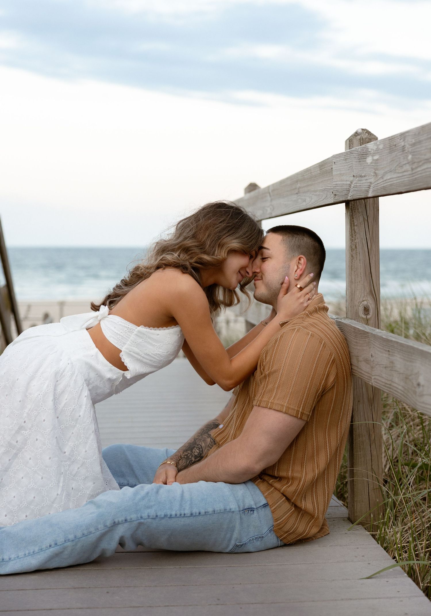 Couple sharing a romantic moment on a beachside boardwalk, woman in white dress leaning toward man in tan shirt.