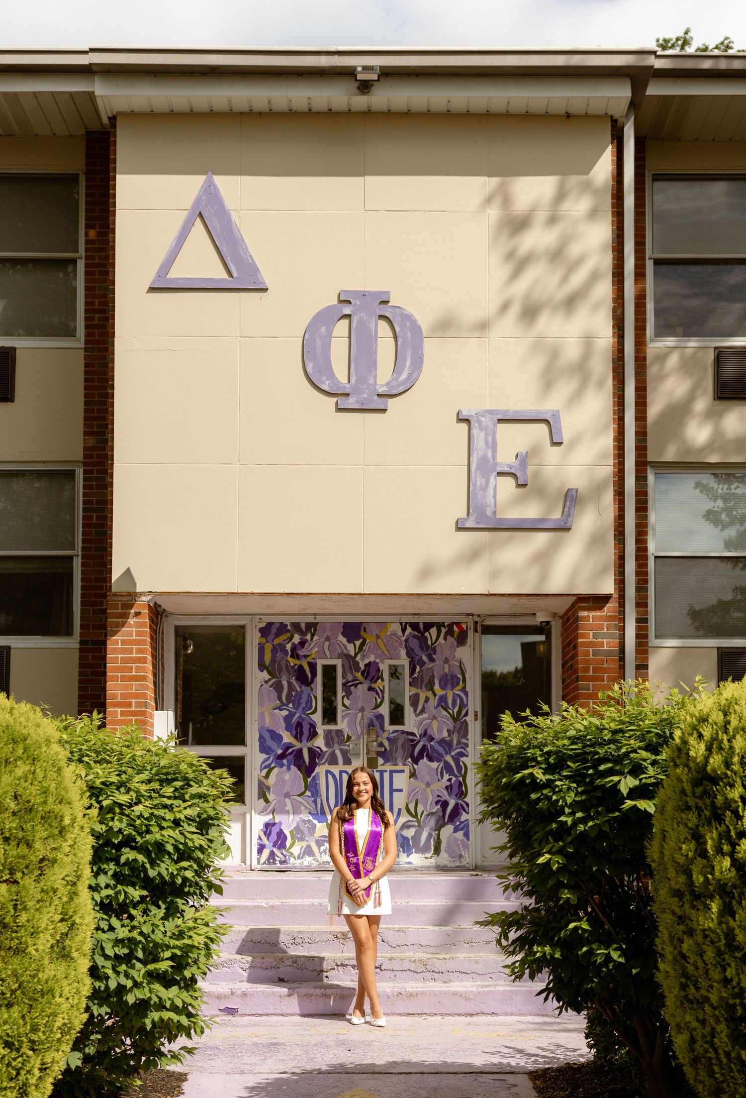 Young woman posing in front of Delta Phi Epsilon sorority house with purple Greek letters and floral door decorations.