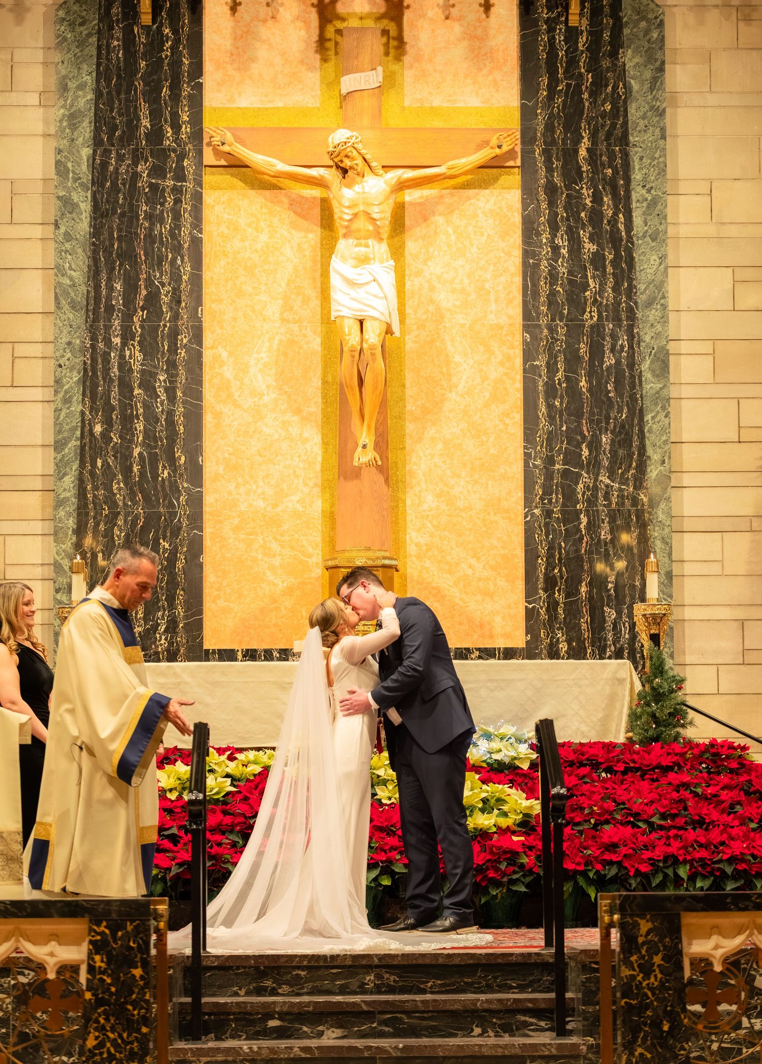 A wedding ceremony takes place in a church with red poinsettias and a large crucifix on the altar.