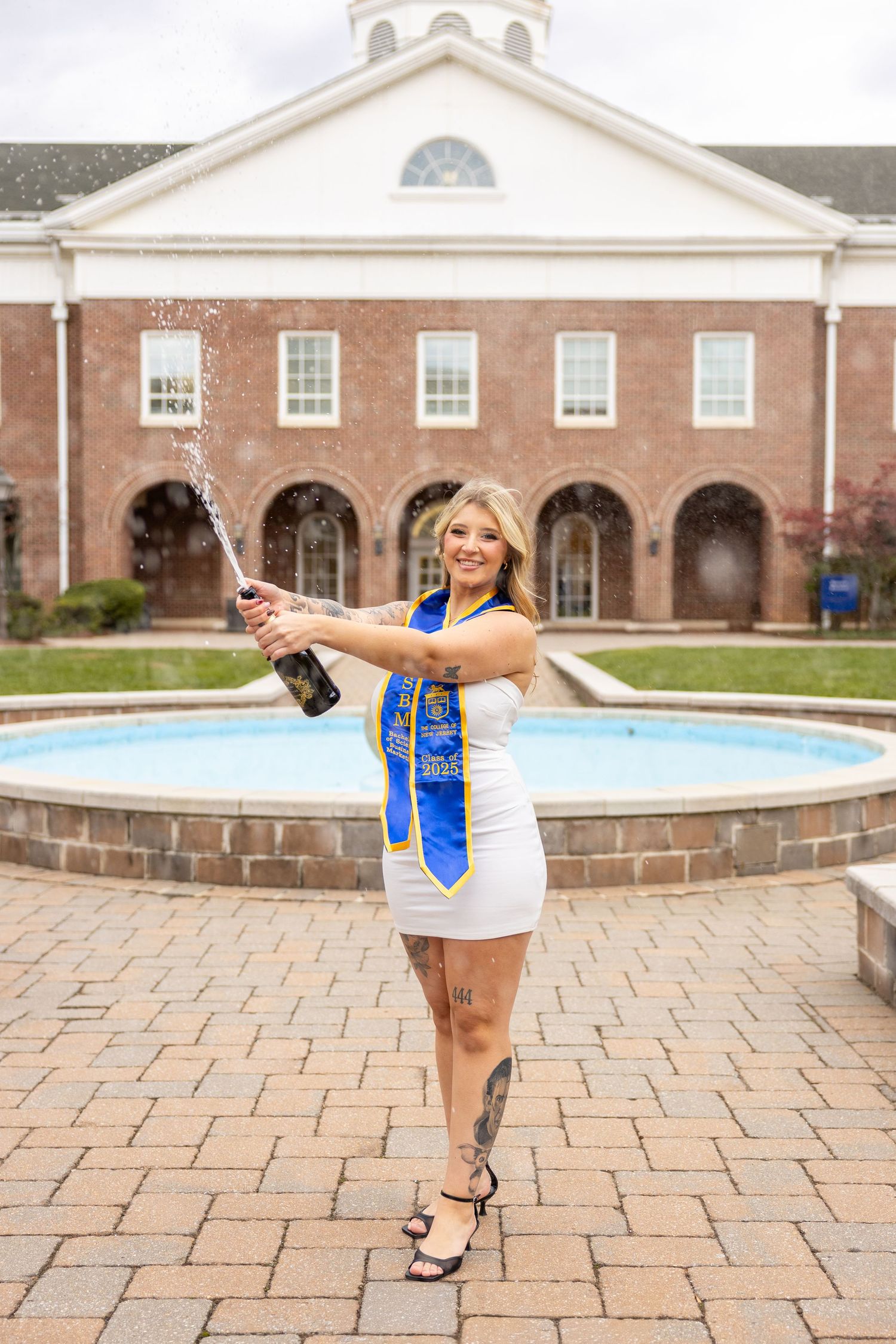 Graduate celebrating with champagne in front of a brick university building with arched doorways and fountain.