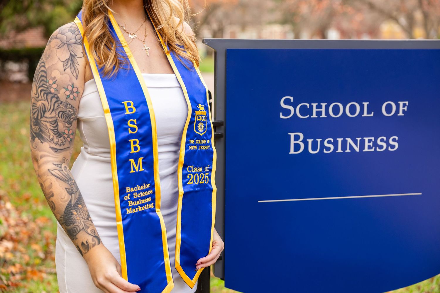 Graduate in cap and gown with tattoos posing next to School of Business sign.