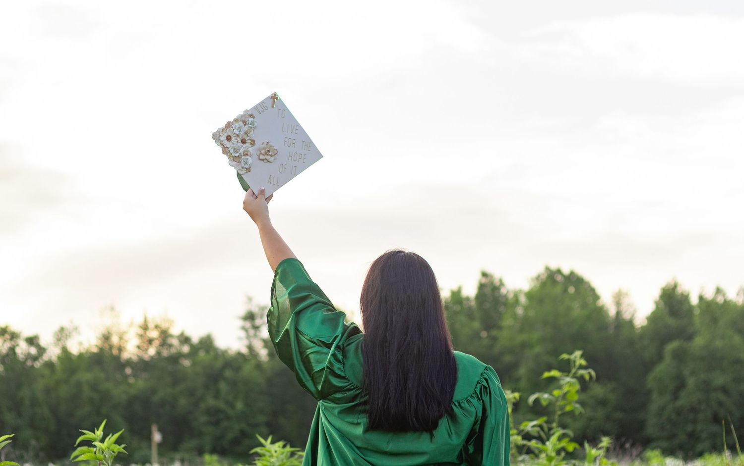 Graduate in green cap and gown poses in front of school sign during sunset celebration.