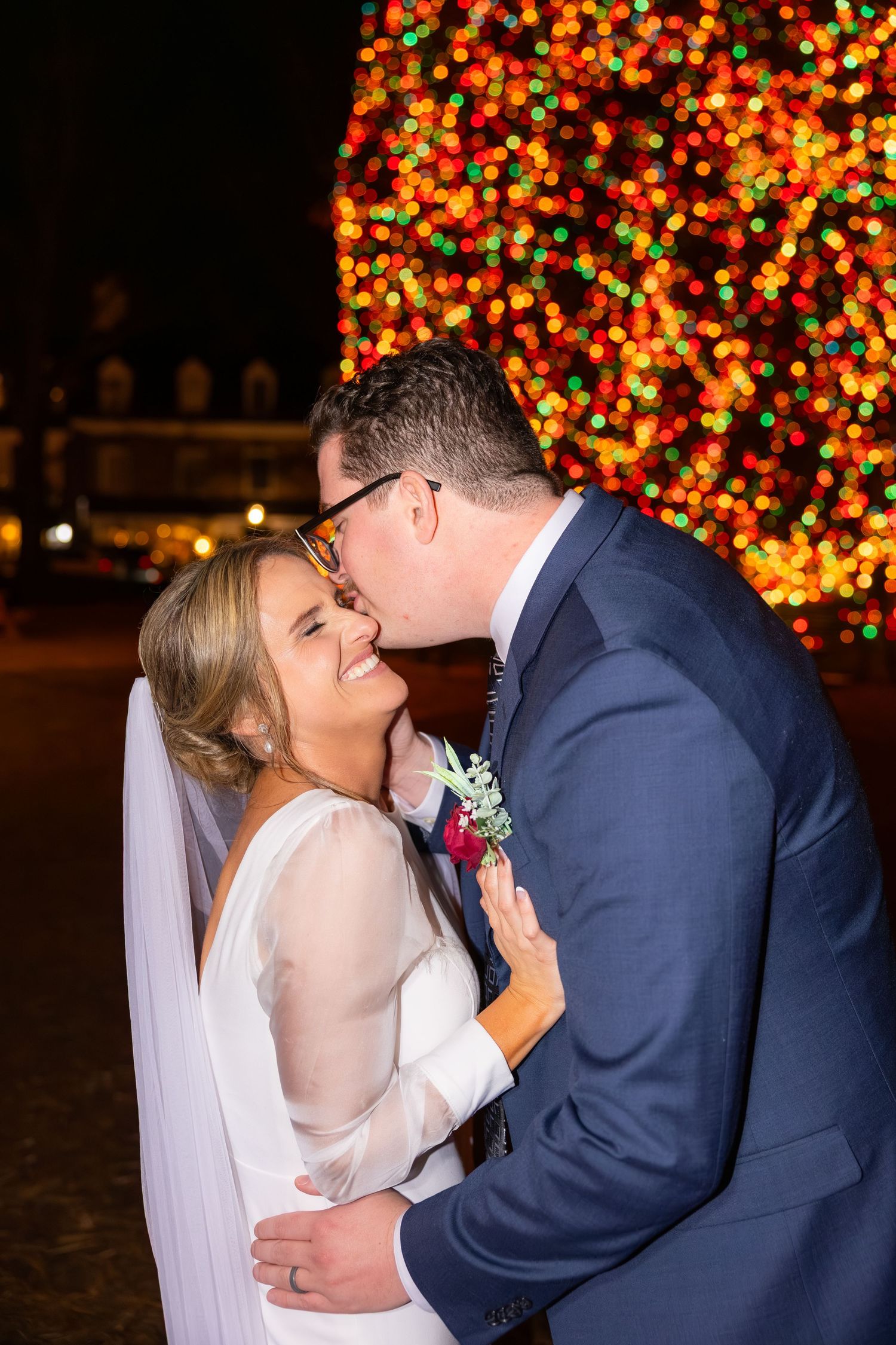 Groom kissing bride on cheek in front of colorful Christmas tree bokeh lights at night wedding celebration.