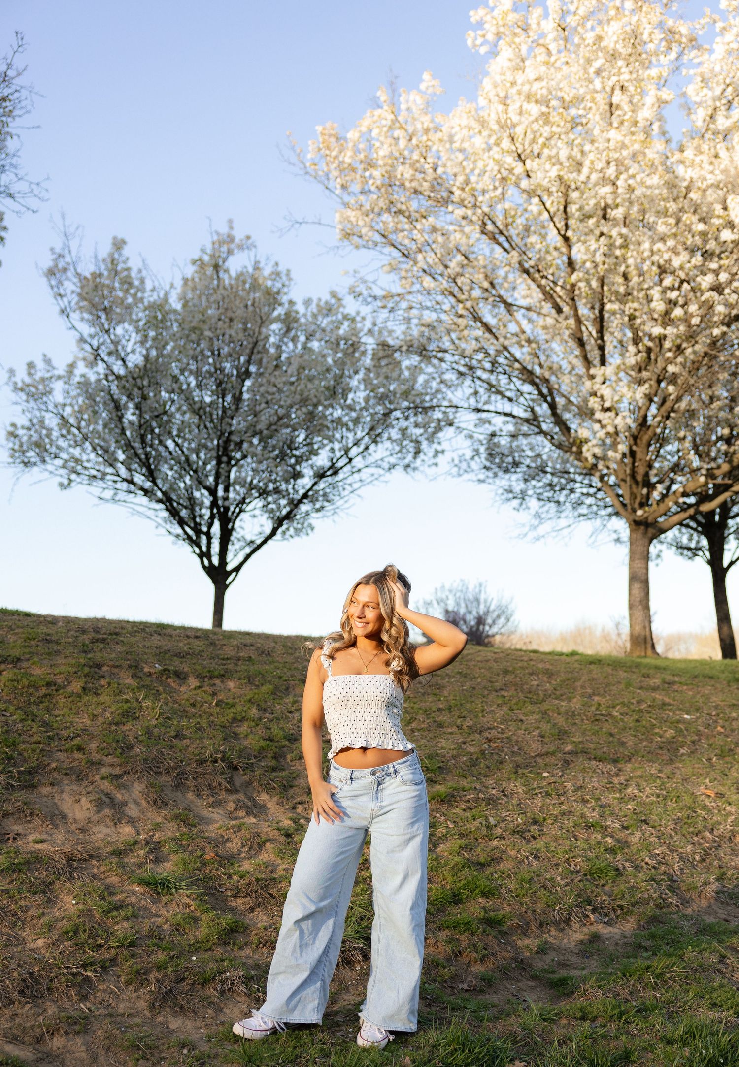 Young woman in crop top and wide-leg jeans poses on grassy hill with blooming white spring trees.