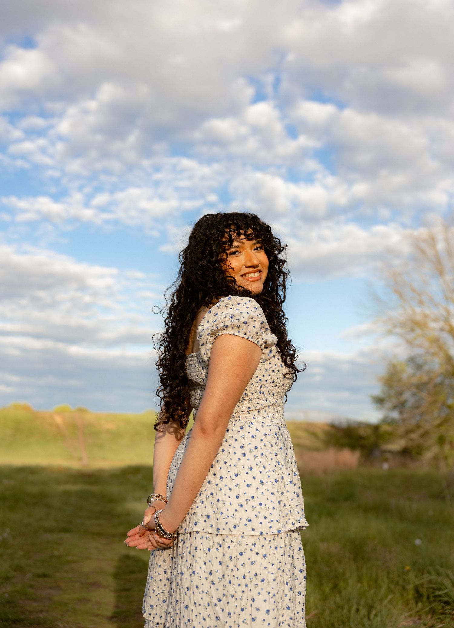 Young woman with curly hair smiling in a floral dress outdoors under a cloudy blue sky.
