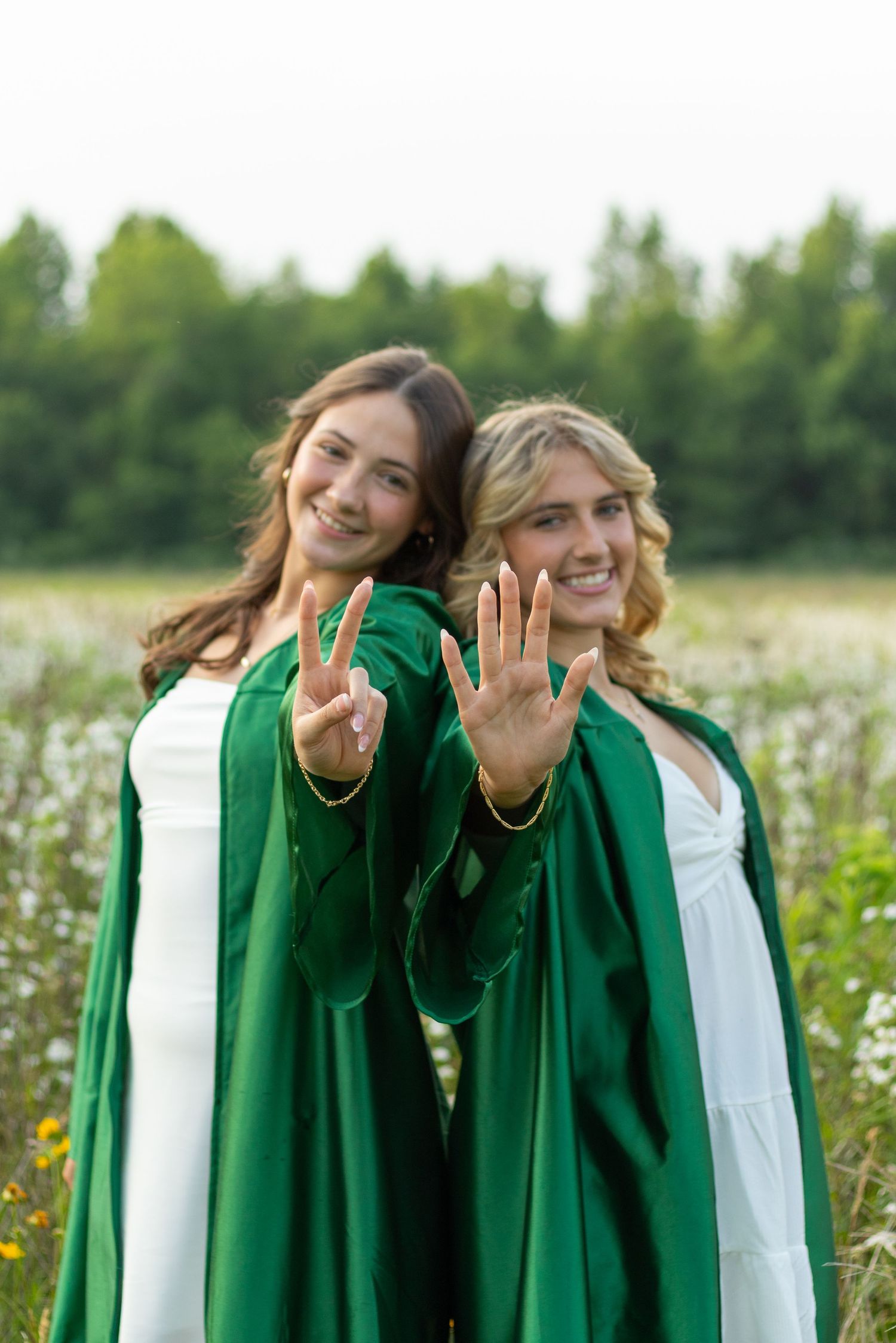 Two graduates in green robes showing peace signs in wildflower field.