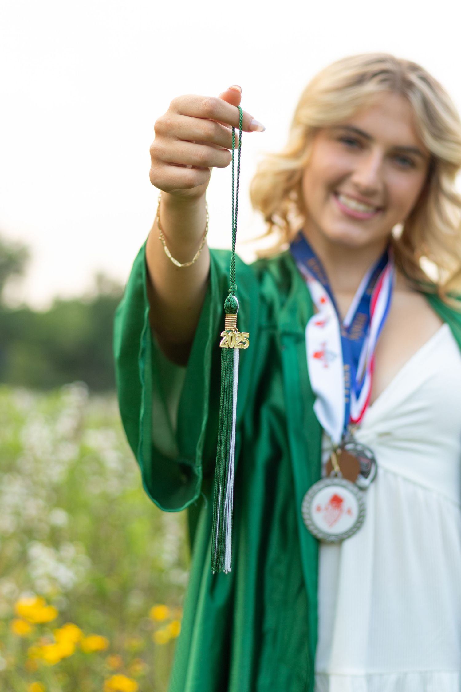 Graduate in green robe displays tassel and medal against natural outdoor backdrop.