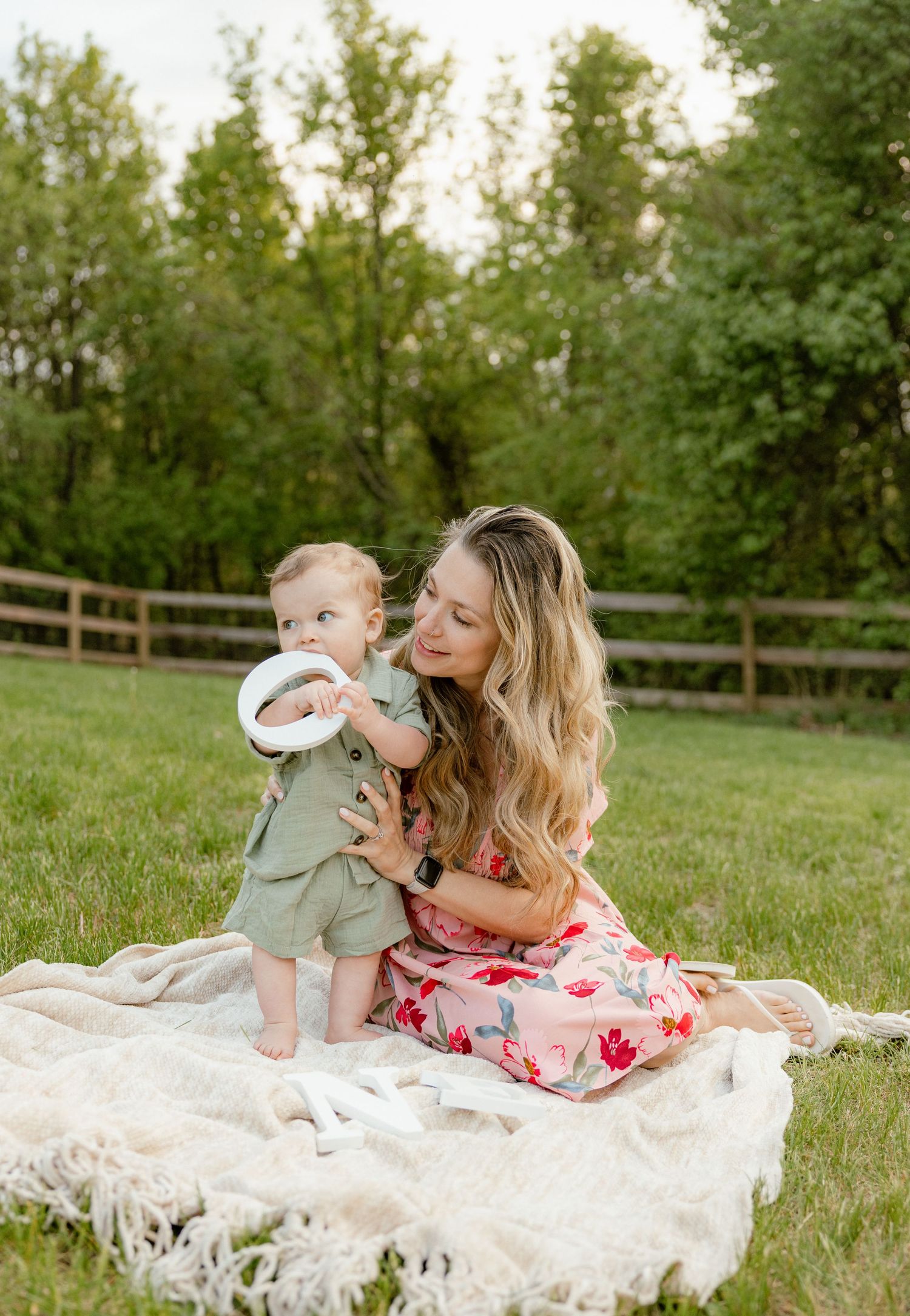 A mother in a floral dress sits on a blanket outdoors with her baby boy in green outfit holding a ring toy.