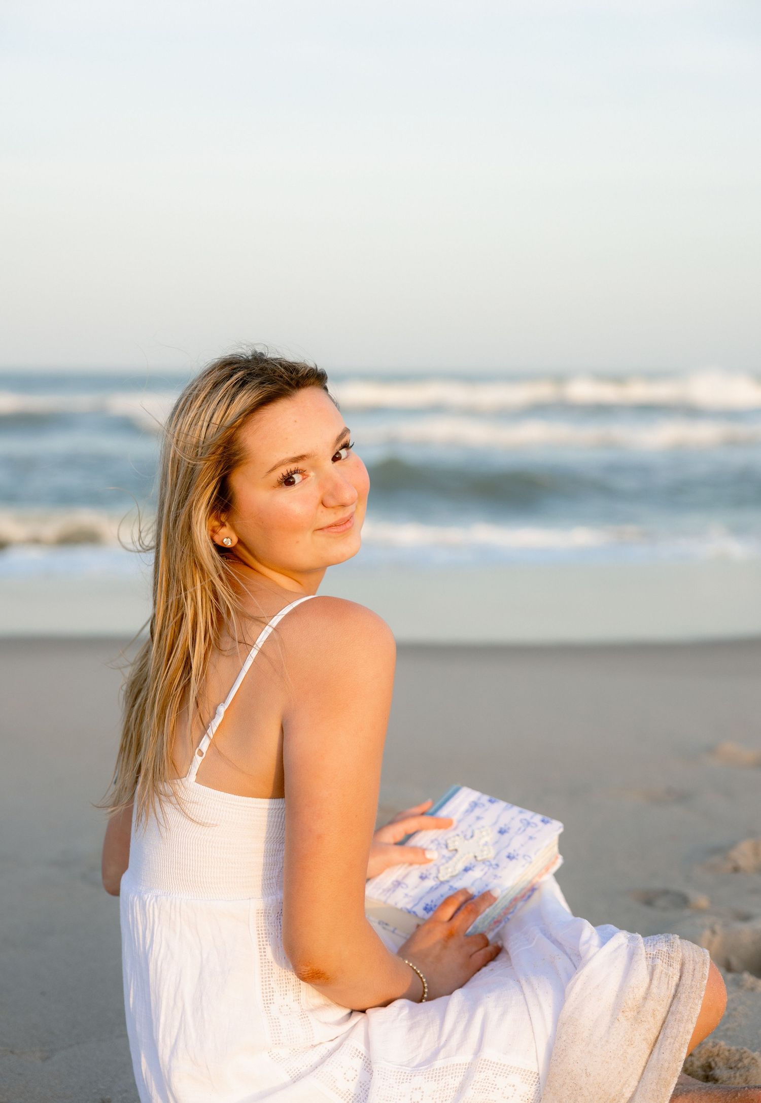 Young blonde woman in white dress sitting on sandy beach holding a book, with ocean waves in the background.