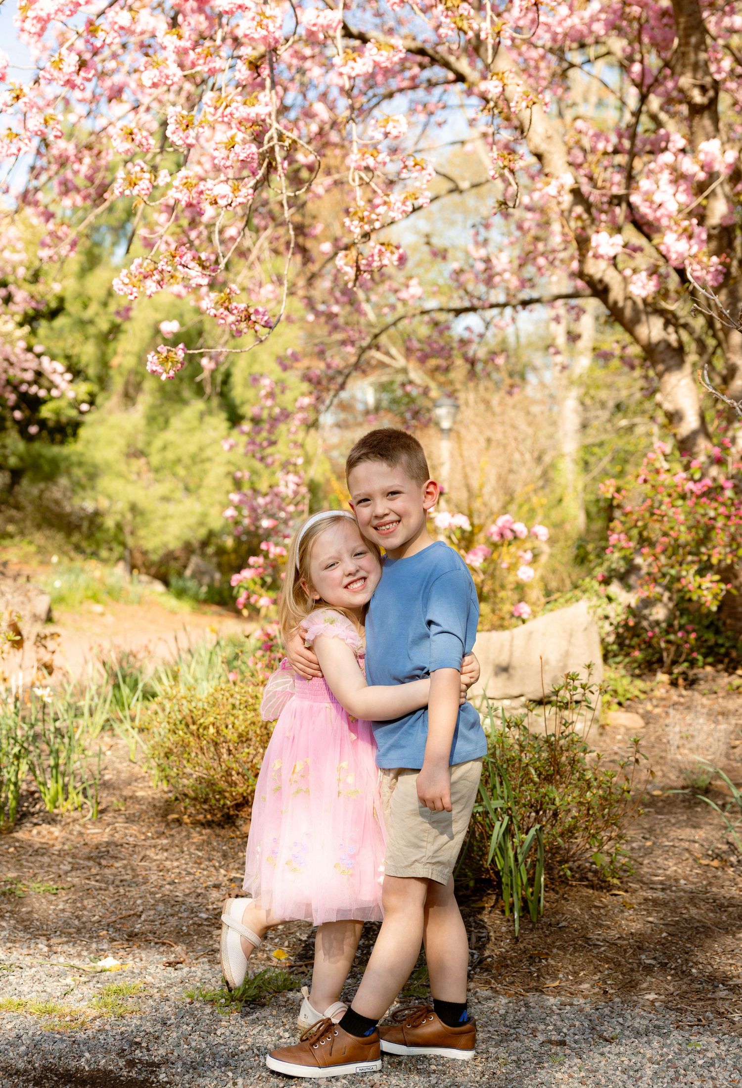 Two children hugging under pink cherry blossom trees in spring, girl in pink dress and boy in blue shirt smiling outdoors.