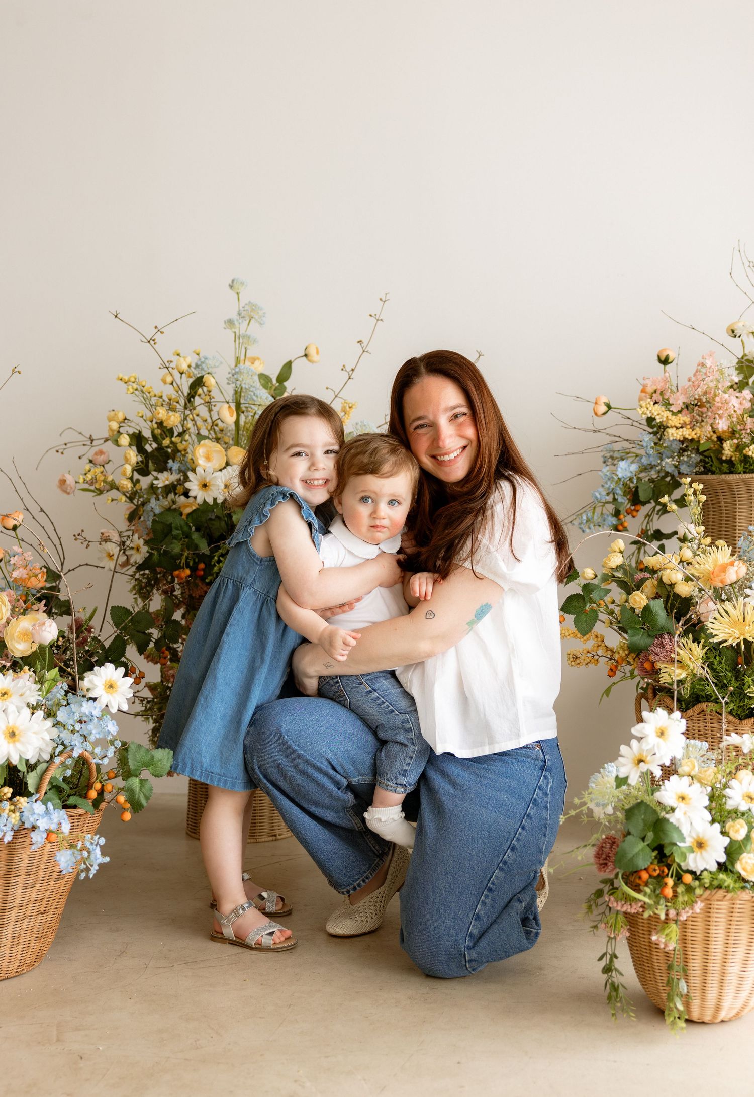 A smiling woman kneels with two young children surrounded by lush floral arrangements in soft yellows and whites.