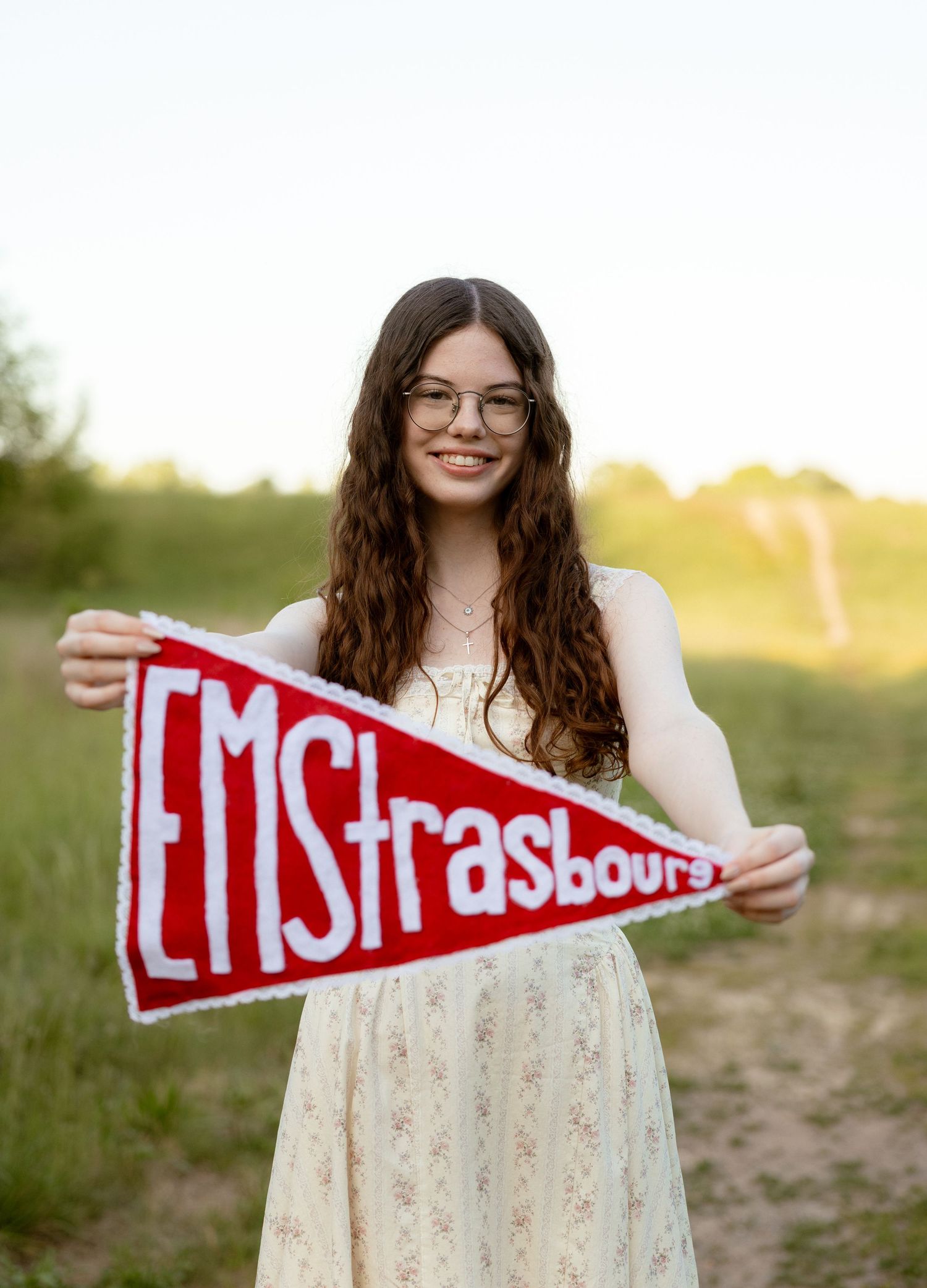 Young woman smiling outdoors holding a red EMStrasbourg pennant flag, wearing glasses and a floral dress.