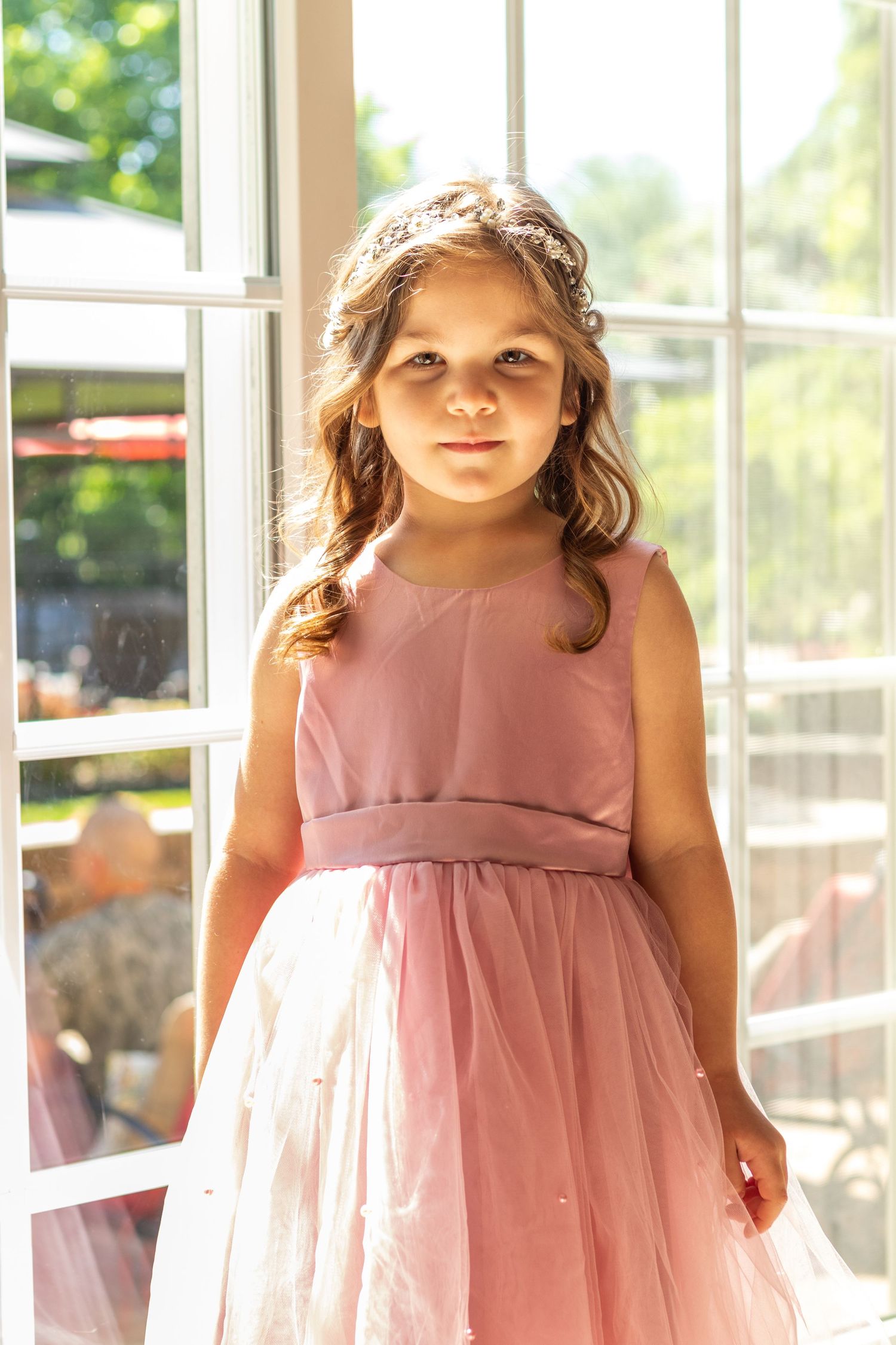 A young attendant in a dusty pink dress stands near sunlit windows.