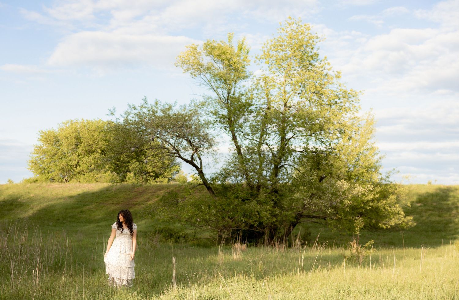 A young woman in a white dress stands in a lush green meadow beside a large tree under a partly cloudy sky.