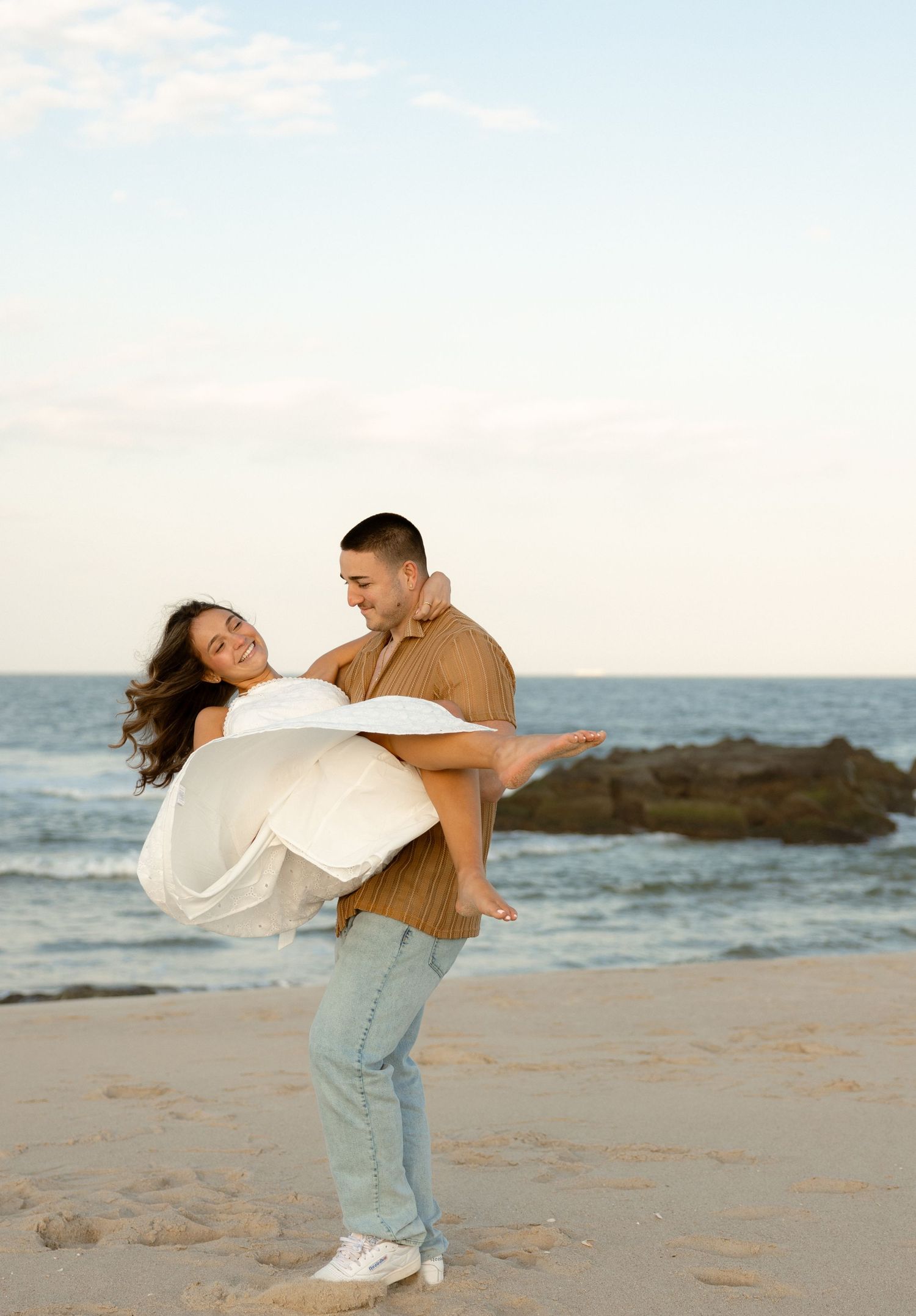 A couple shares a romantic moment on the beach, as a man lifts a woman in a white dress near the ocean.