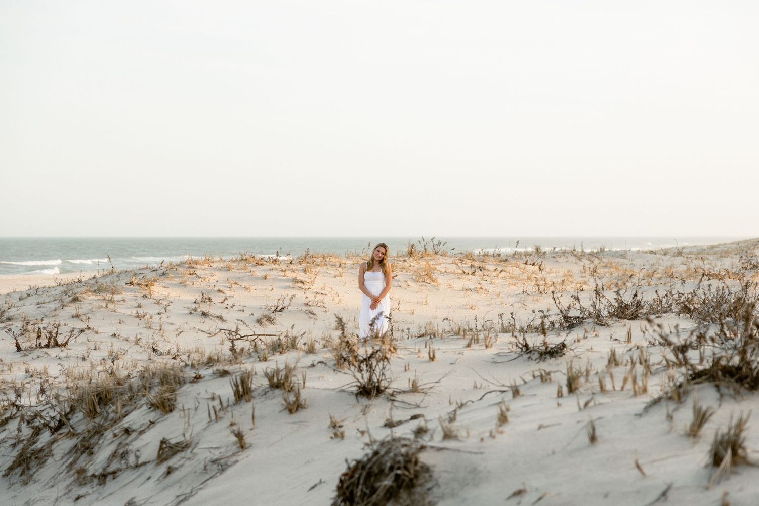 A woman in a white dress stands on sandy beach dunes with ocean waves in the background on an overcast day.