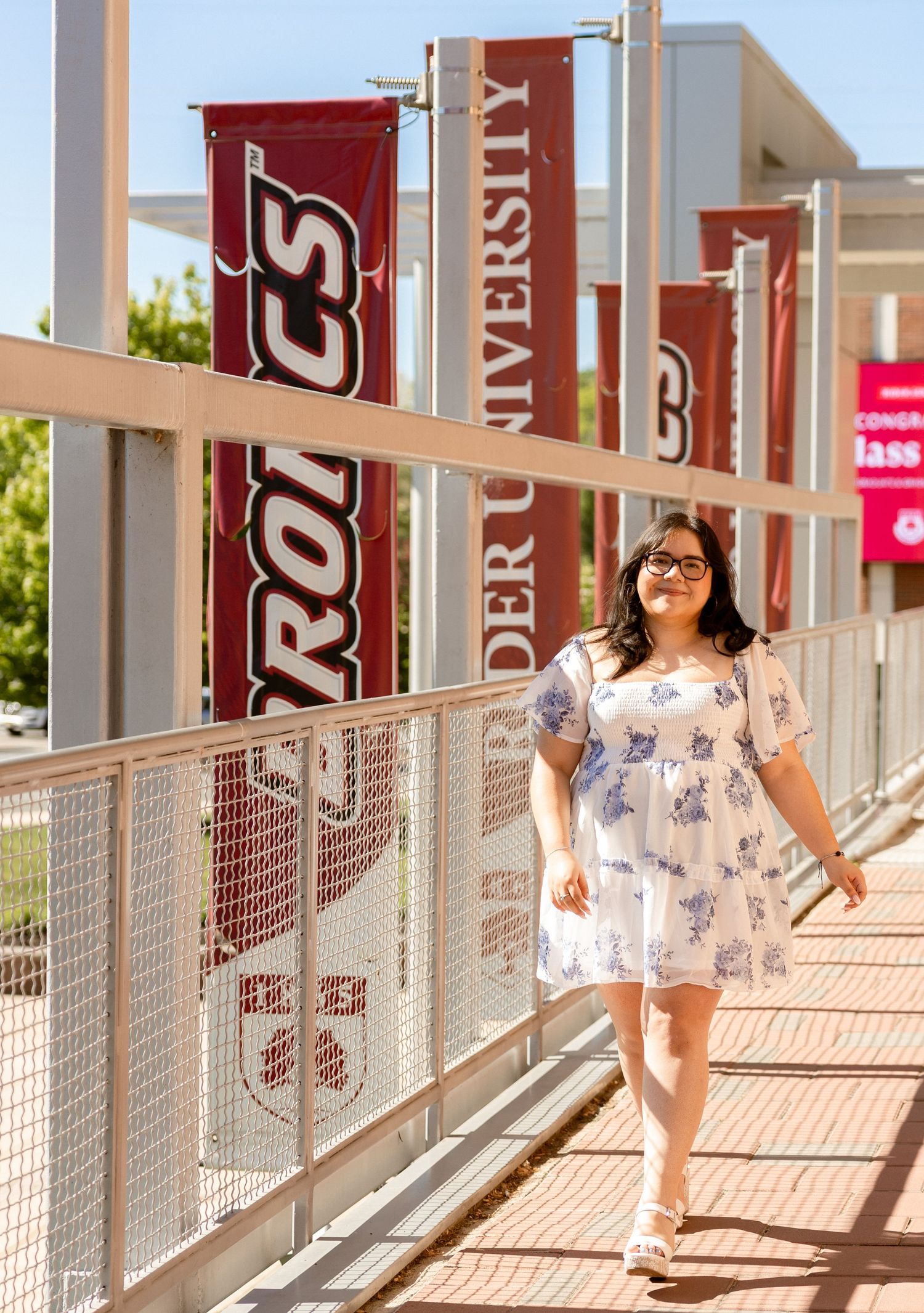 A student walks along a bridge at Boulder University, with red Broncos banners lining the path on a sunny day.