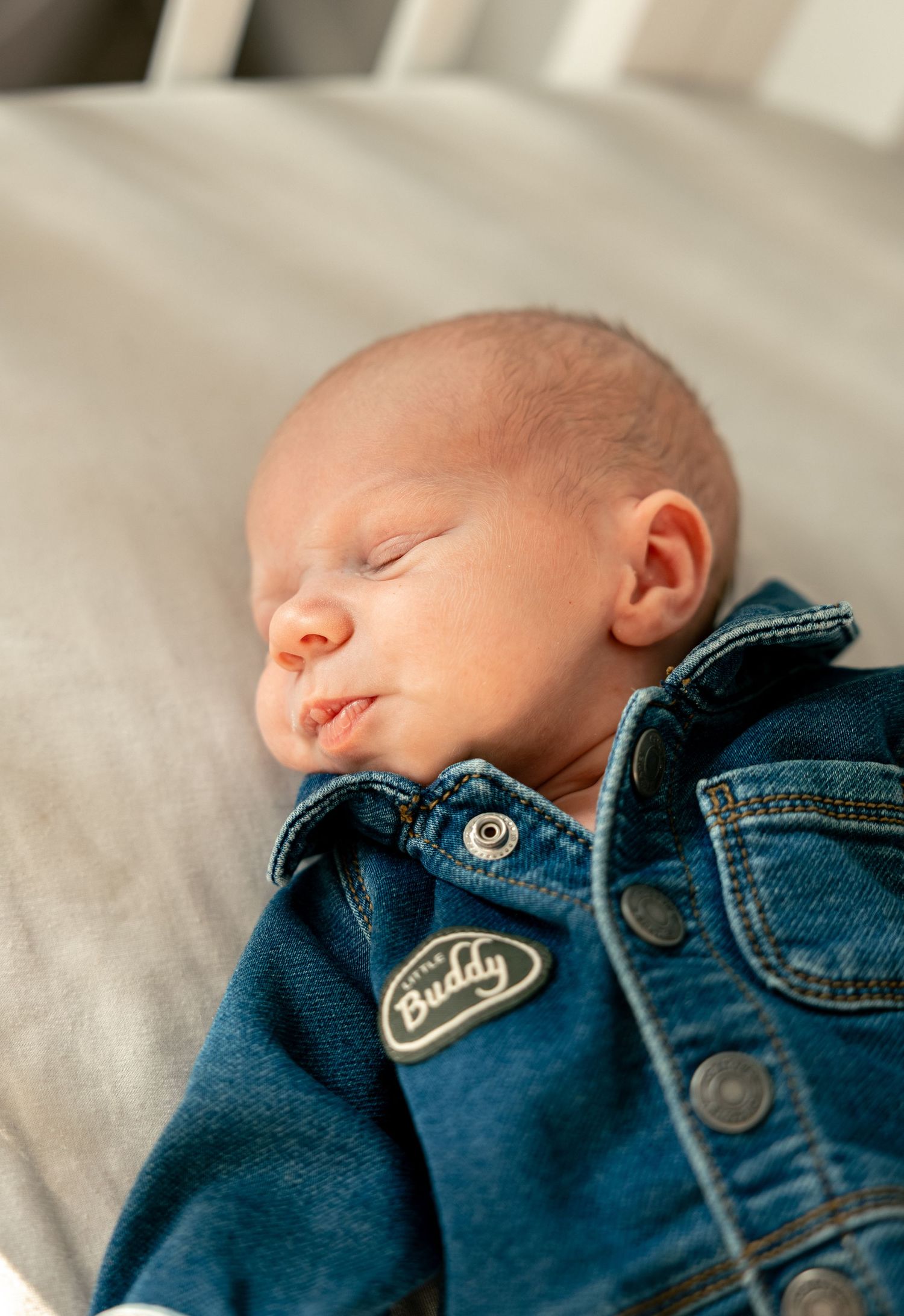 Sleeping newborn baby wearing a denim jacket with a Buddy patch, resting peacefully on a gray surface.