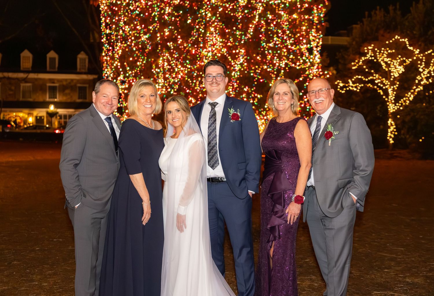 Wedding party poses outdoors against beautifully lit trees during evening celebration.