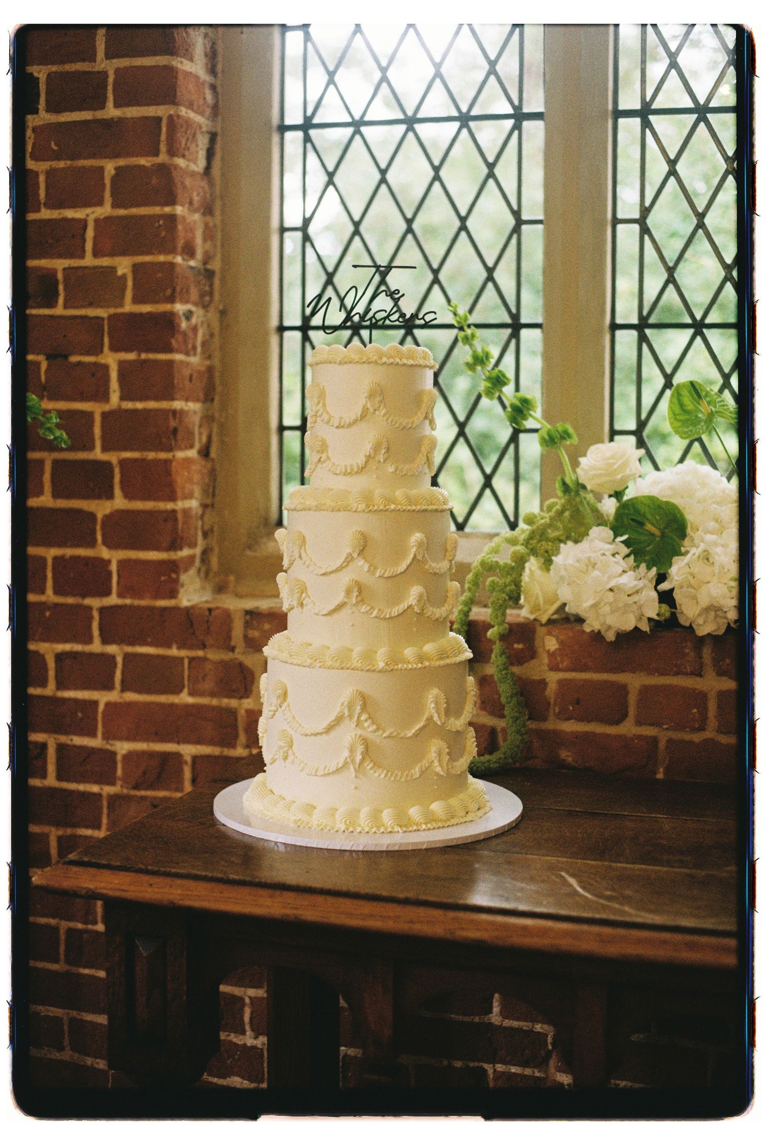 Elegant five-tier white wedding cake with ruffle details displayed on wooden table before leaded glass windows.