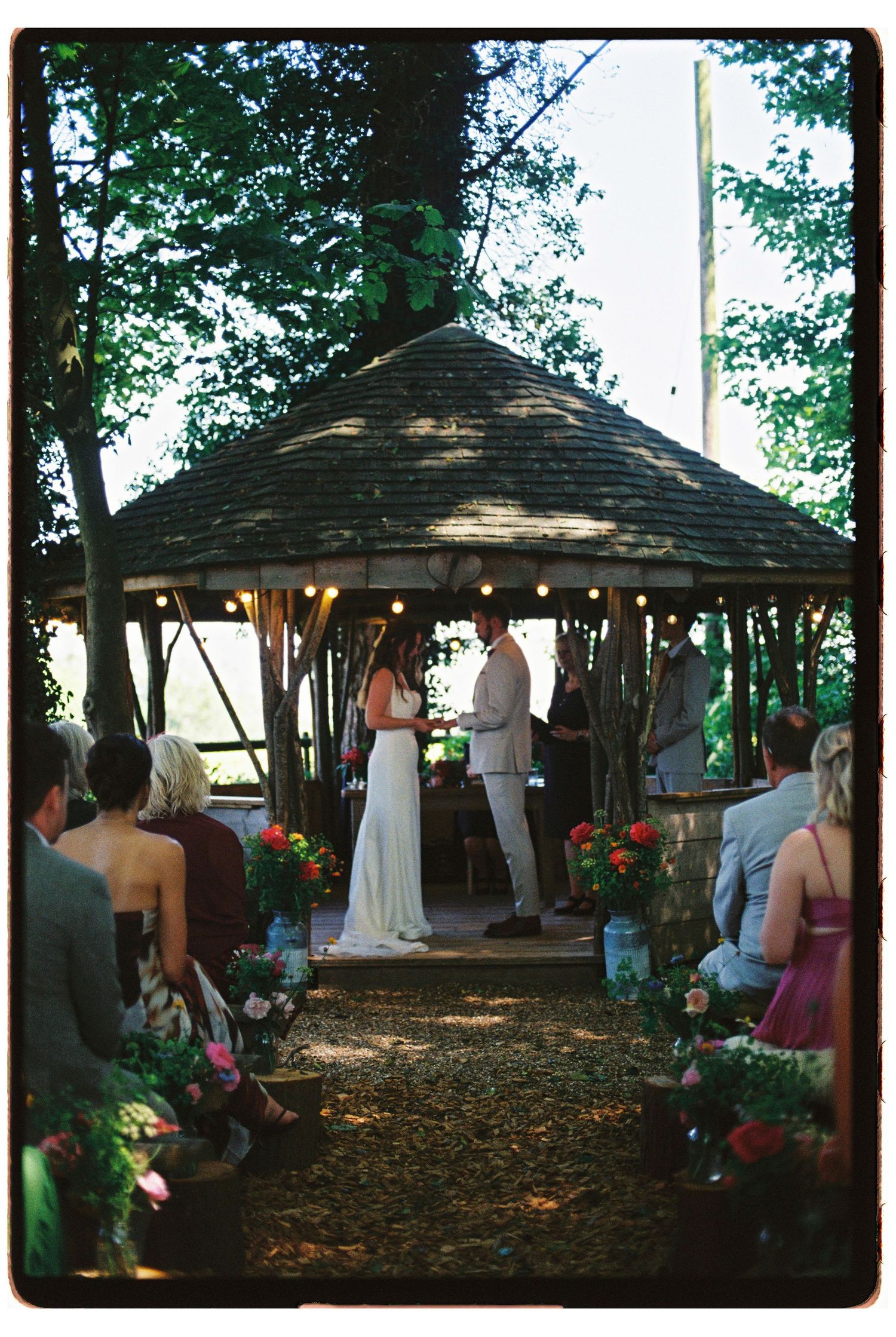Outdoor wedding ceremony under a gazebo adorned with string lights and floral arrangements in a forest setting.