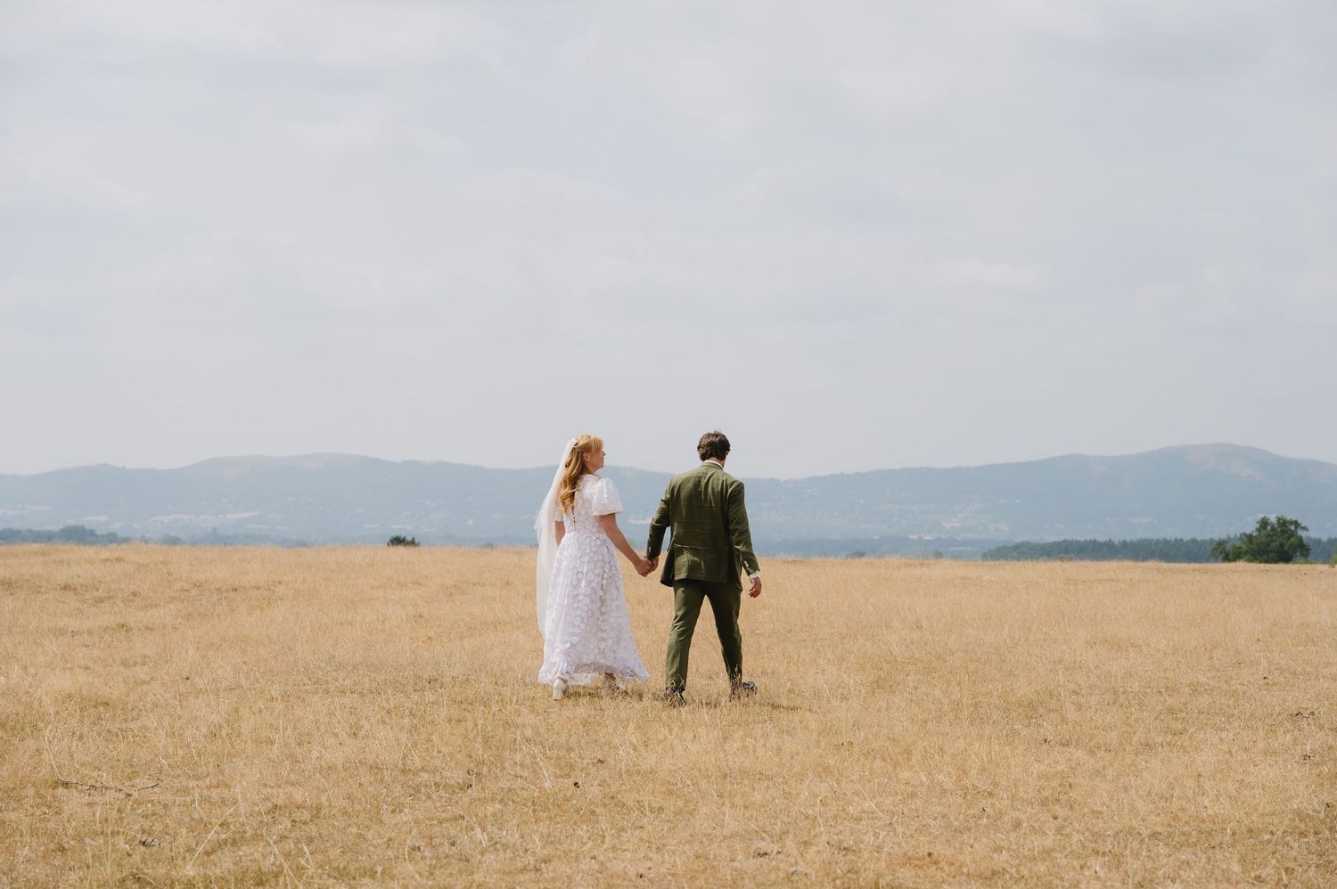 Bride and groom holding hands walking through a vast golden wheat field on their wedding day.