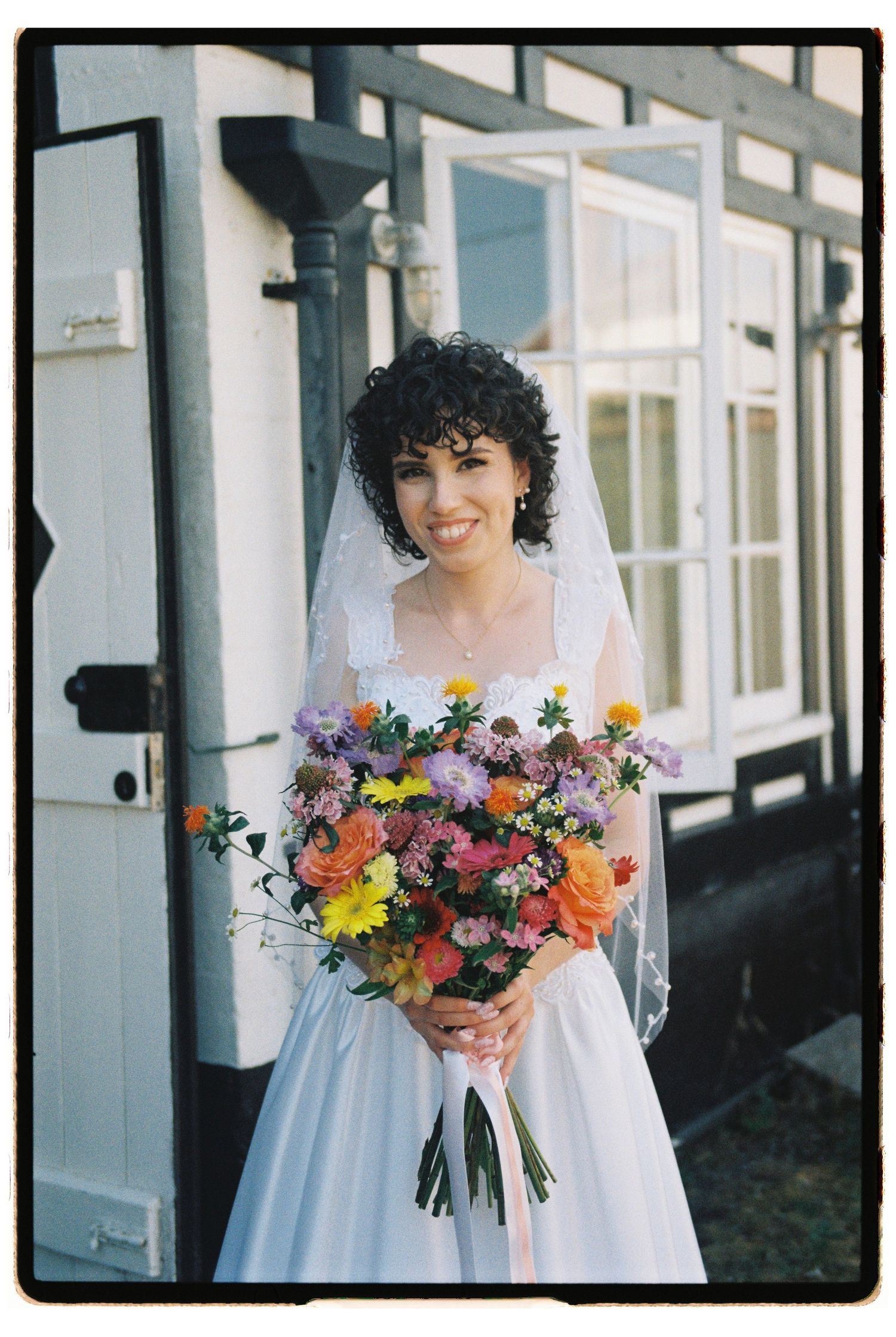 Smiling bride in white gown and veil holds colorful flower bouquet outside a white building.
