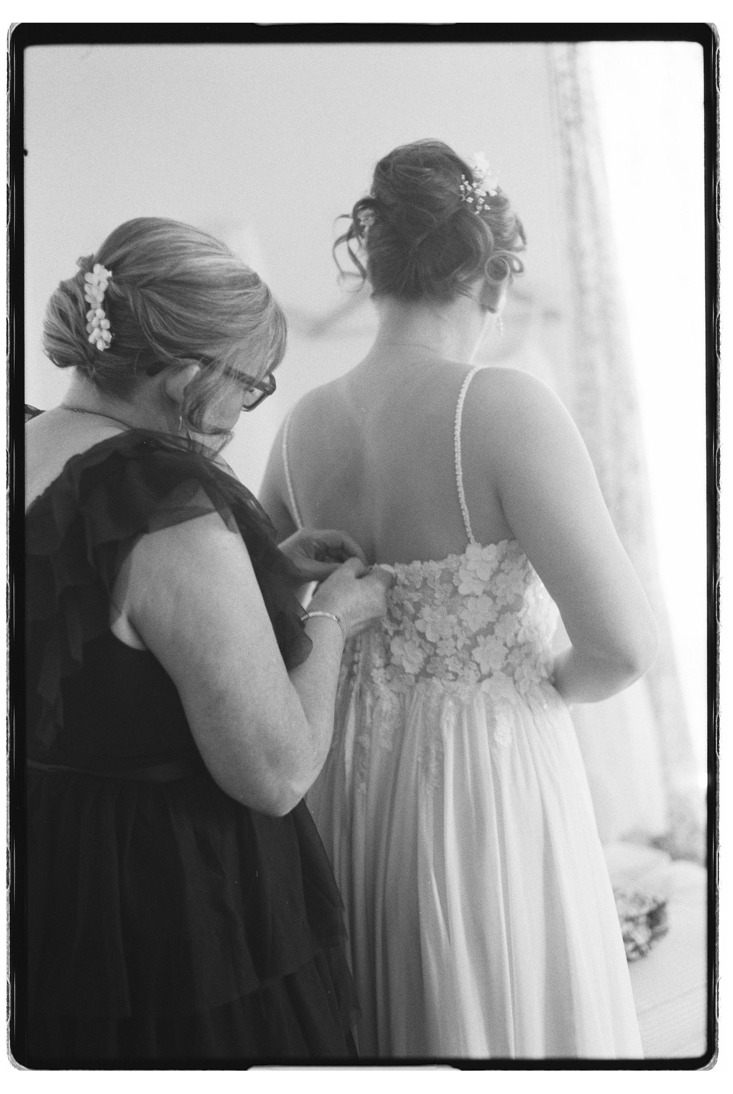 Black and white film photo of a woman fastening the back of a brides wedding dress before the ceremony.