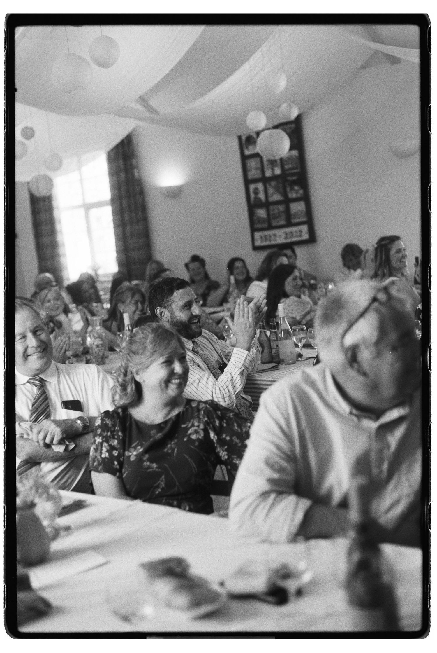 Black and white film photo of smiling wedding guests seated at tables during an indoor wedding reception.
