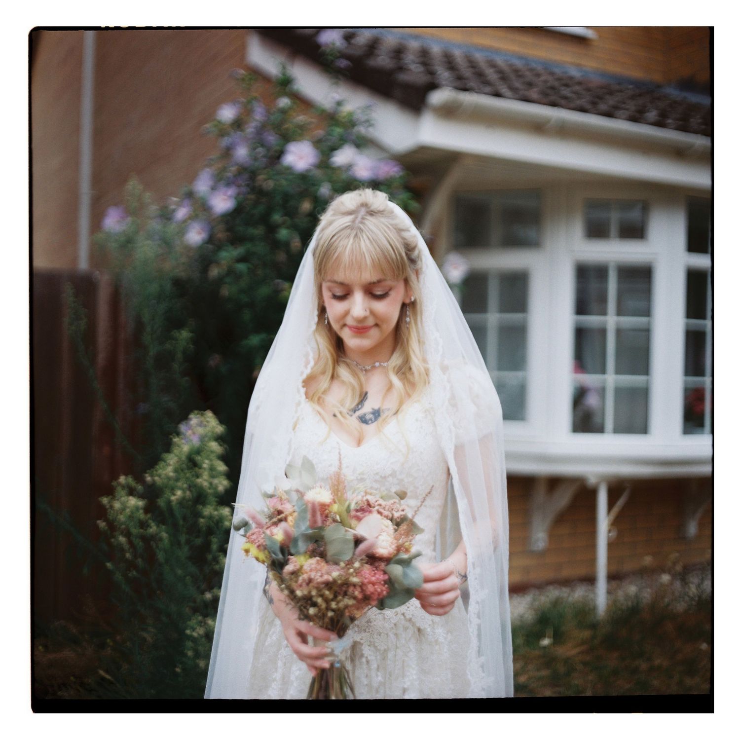 Blonde bride in lace dress holds floral bouquet outside home before wedding ceremony.