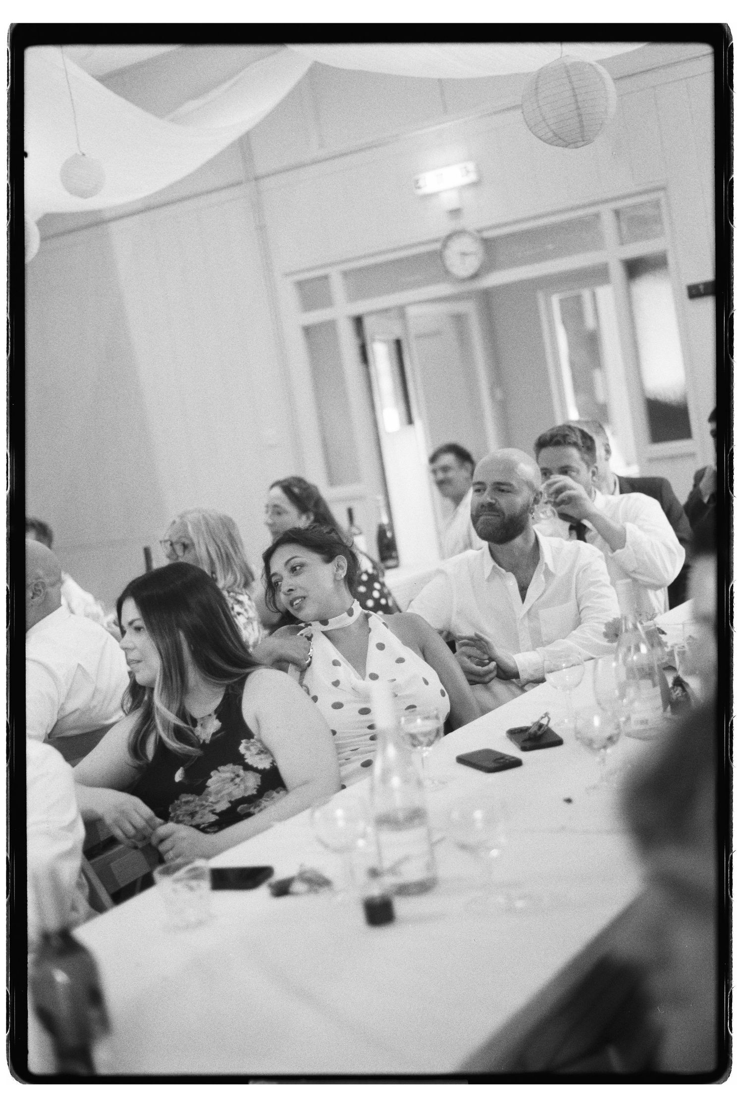 Black and white film photo of wedding guests seated at a long table during an indoor wedding reception.