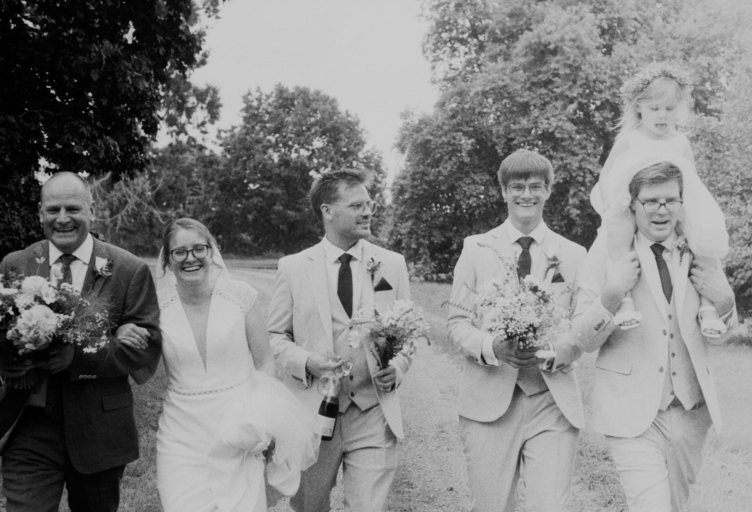 Black and white photo of happy wedding party walking together outdoors, including bride, groom, and guests.