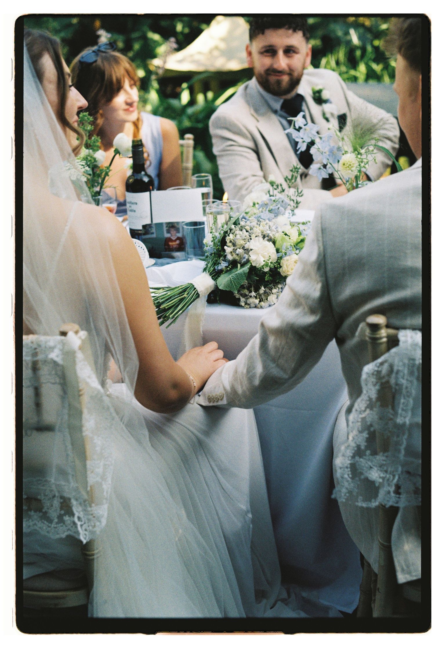 Bride and groom holding hands at outdoor wedding reception table with white floral bouquet.