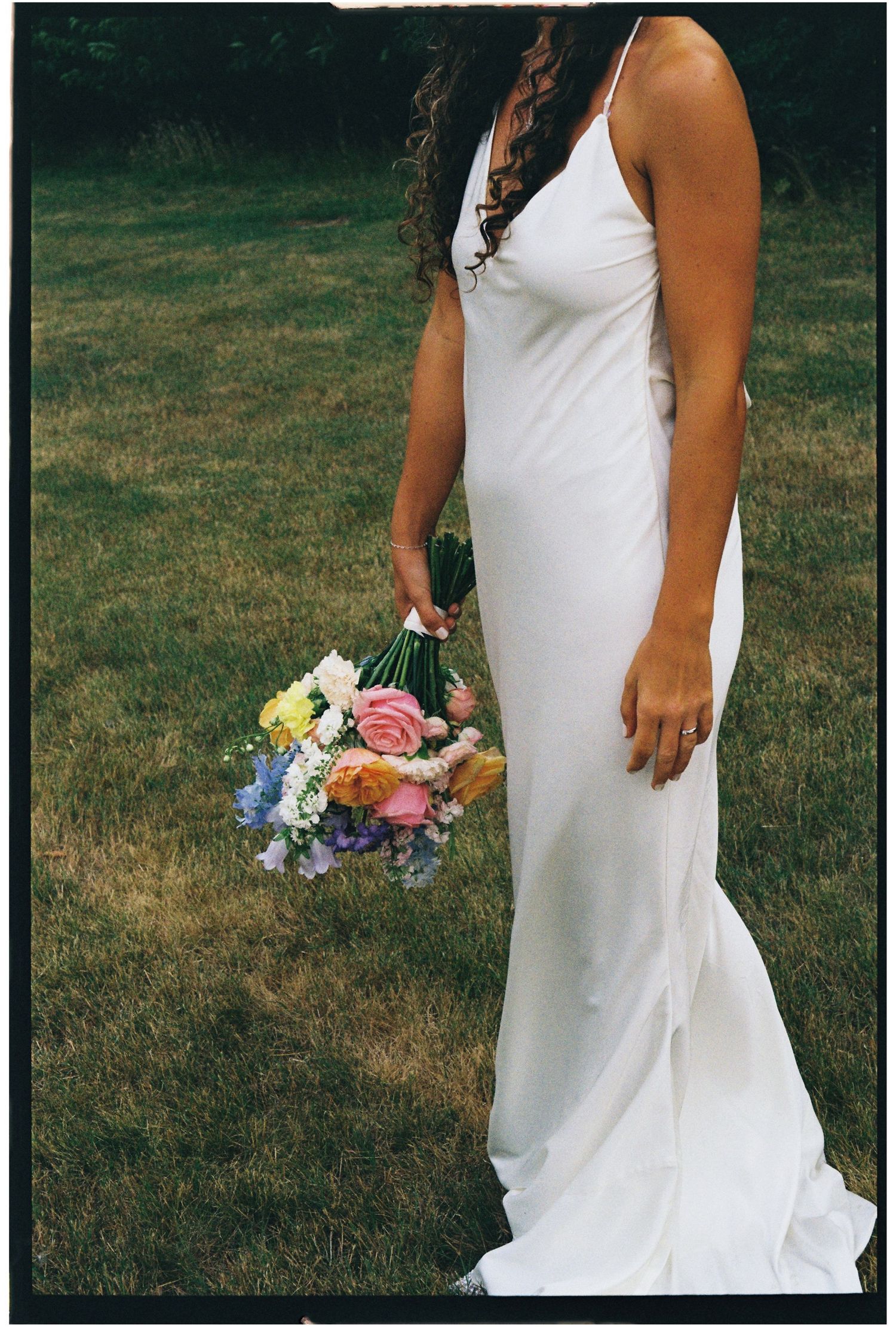 Bride in white slip gown holds colorful wildflower bouquet on green lawn.