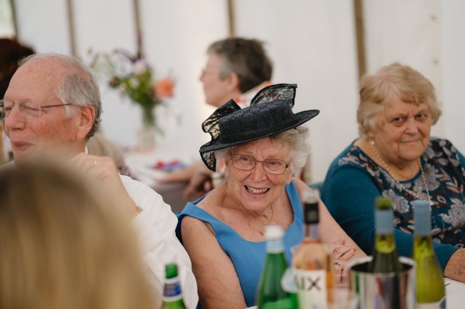 Smiling elderly woman in a navy fascinator hat enjoying a wedding reception in a marquee.
